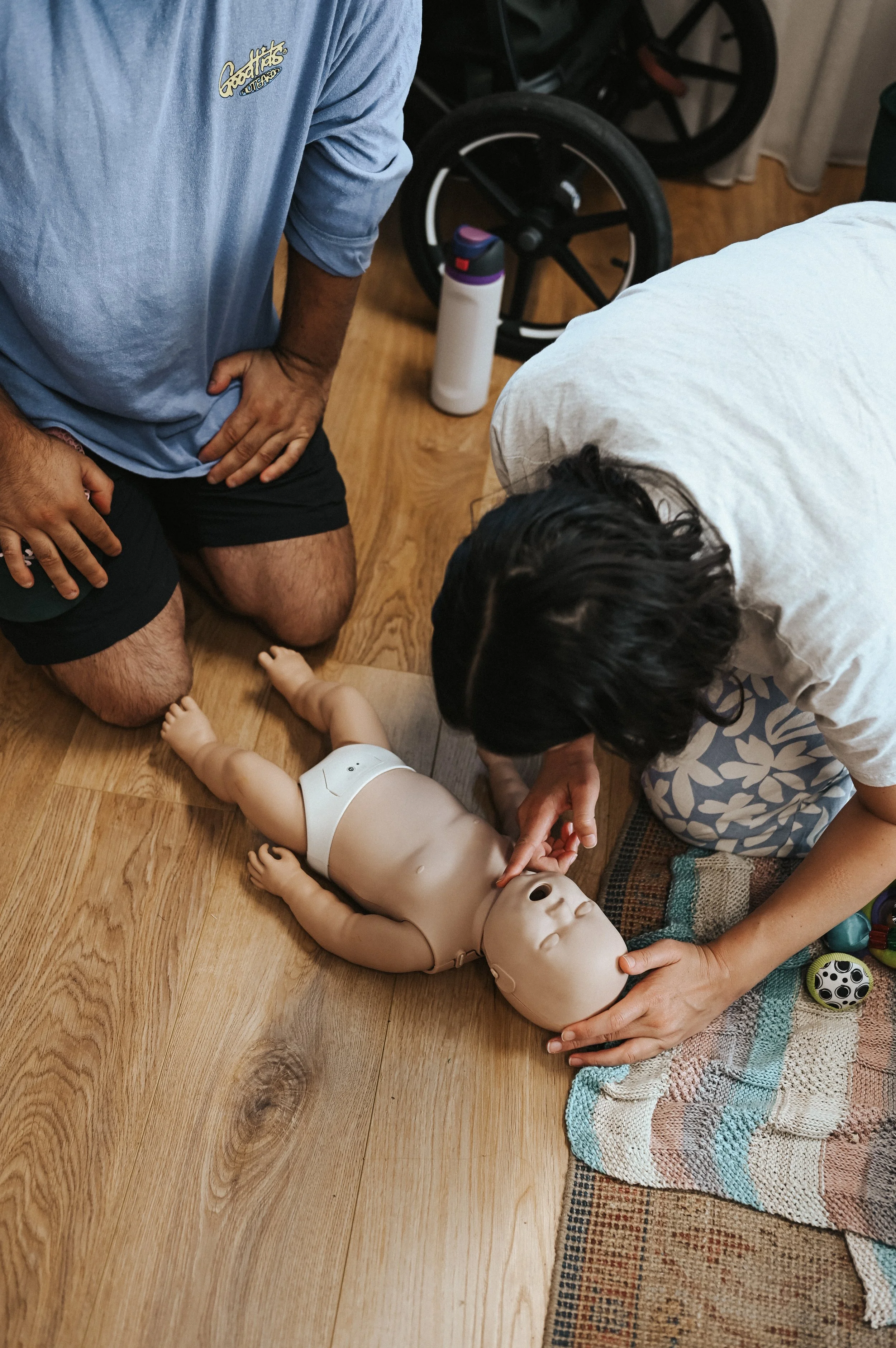 A person kneeling on the floor performing CPR on a doll, with another person assisting.