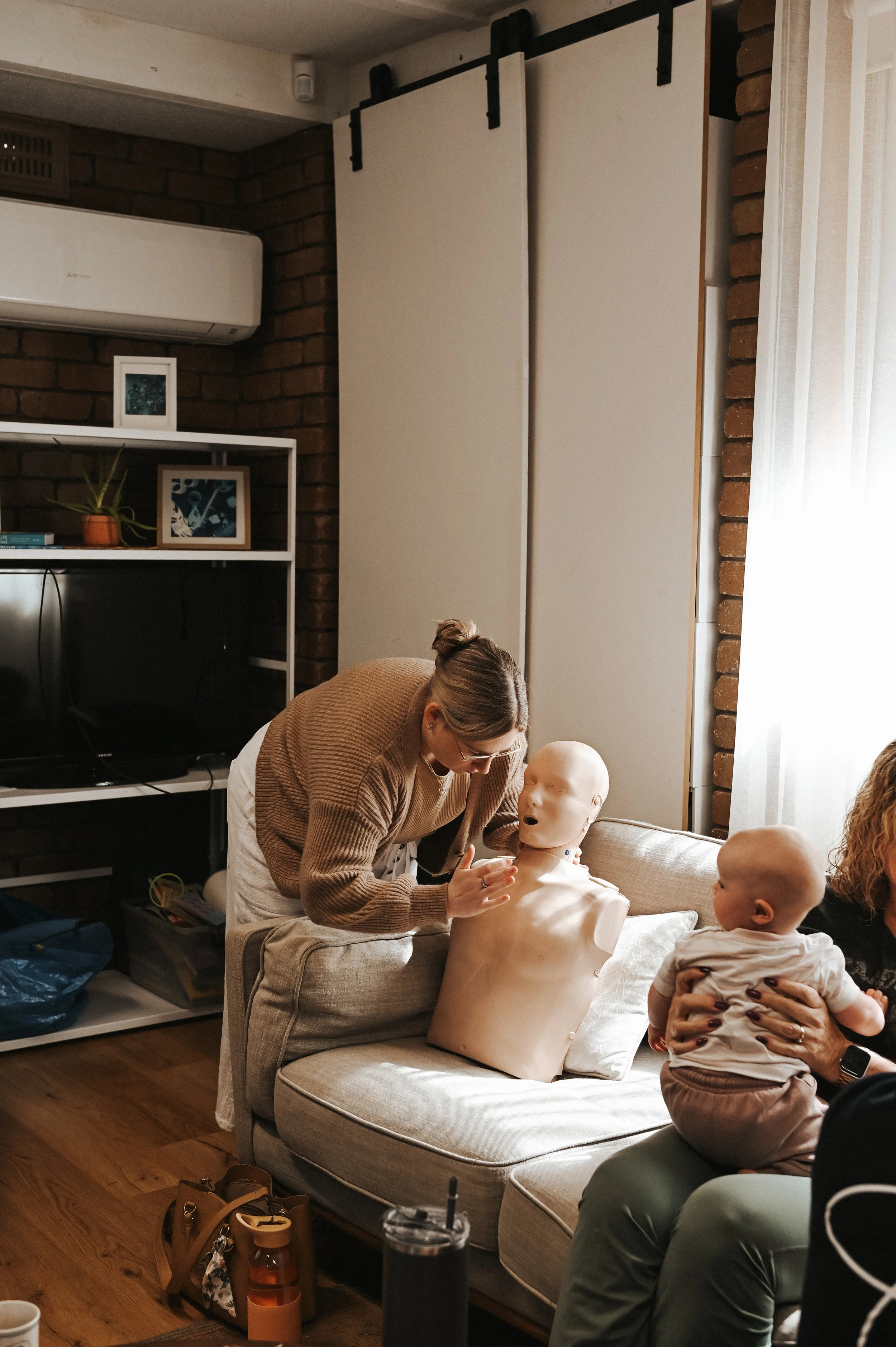 A woman demonstrating CPR on a medical training mannequin with a baby girl sitting nearby during a demonstration or class in a cozy living room.
