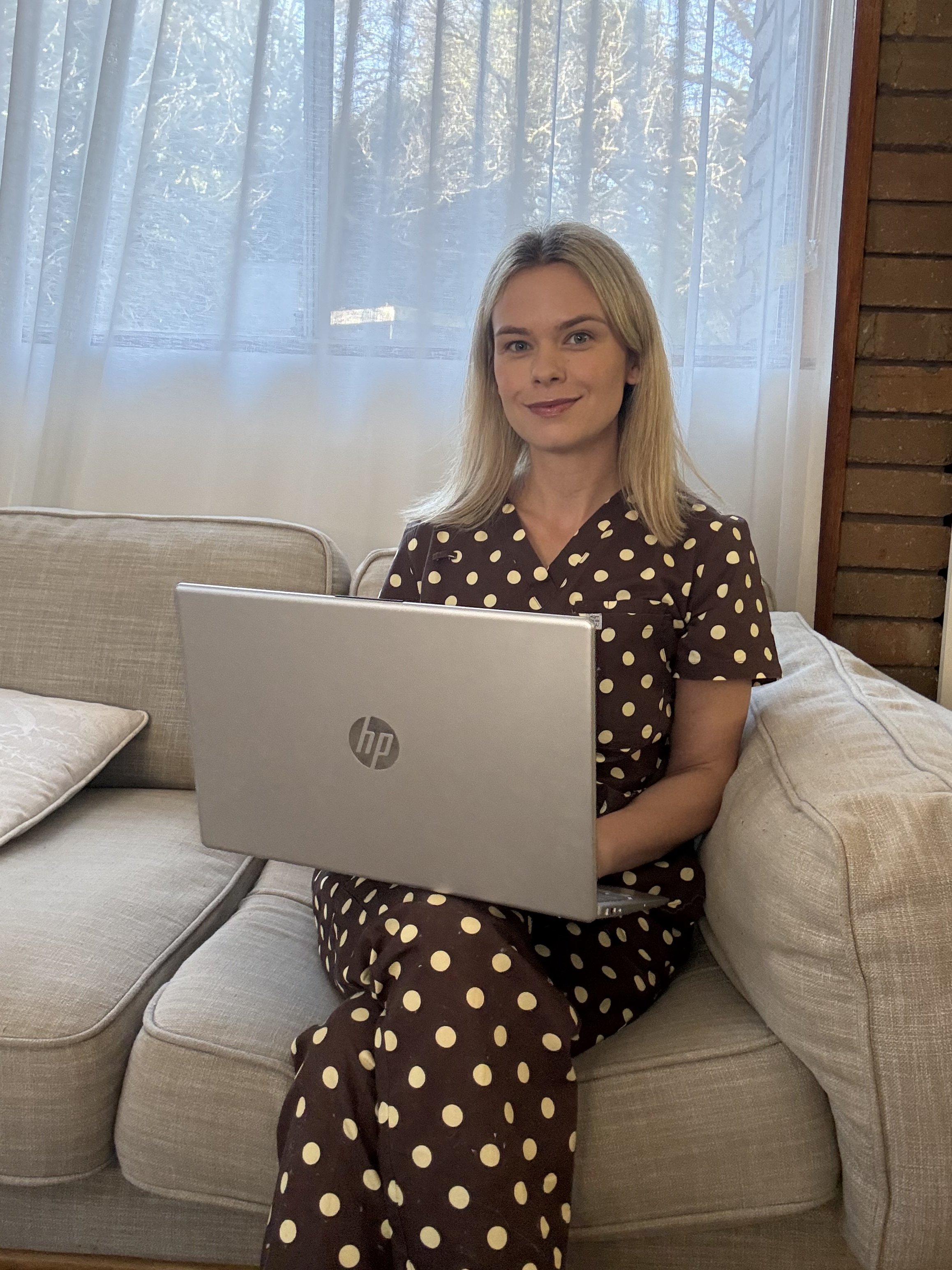 A woman sitting on a beige sofa, using a silver HP laptop, wearing brown scrubs with yellow polka dots, in a room with large window and a brick wall.