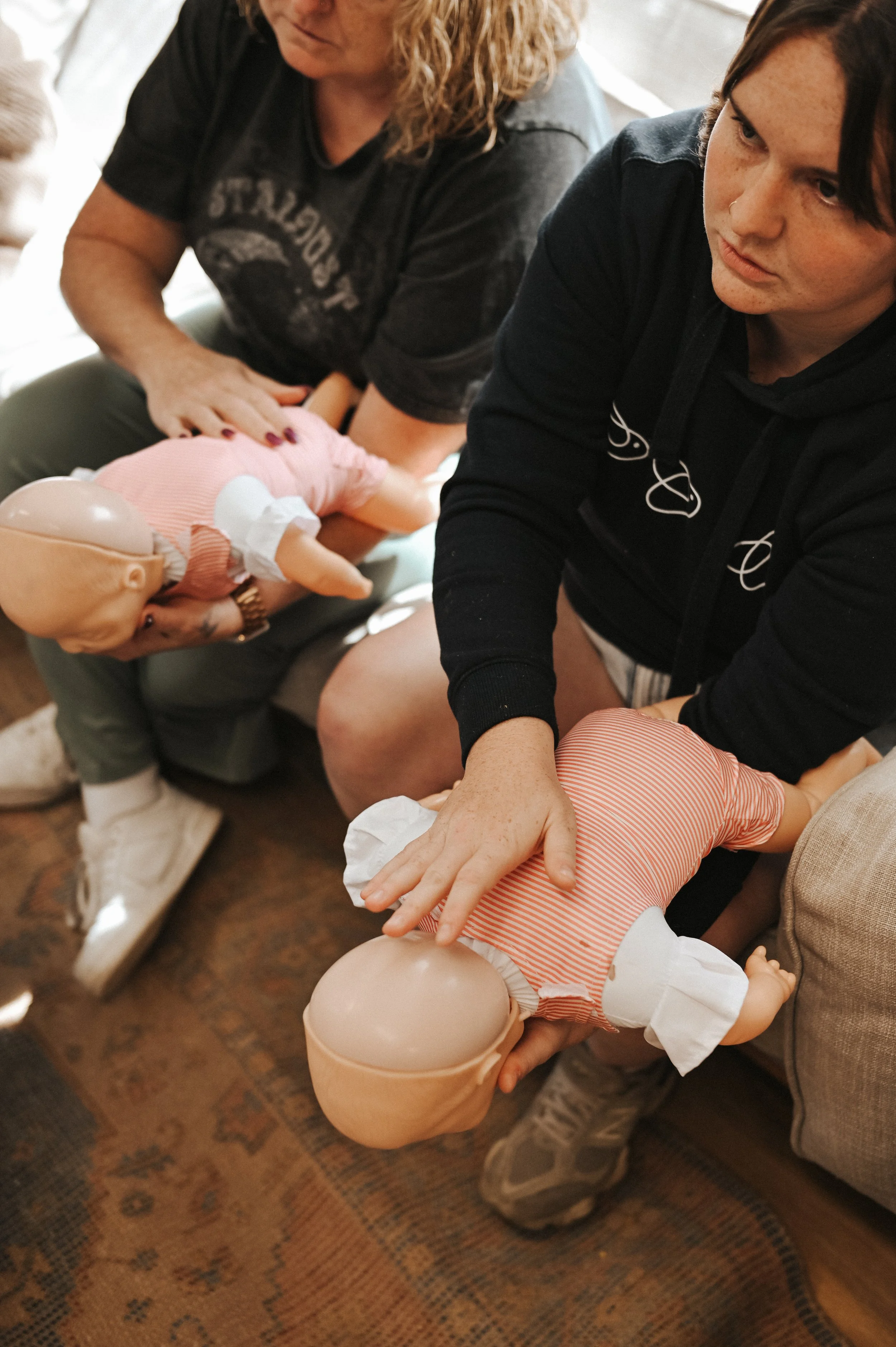 Two women practicing CPR on infant mannequins in an indoor setting.