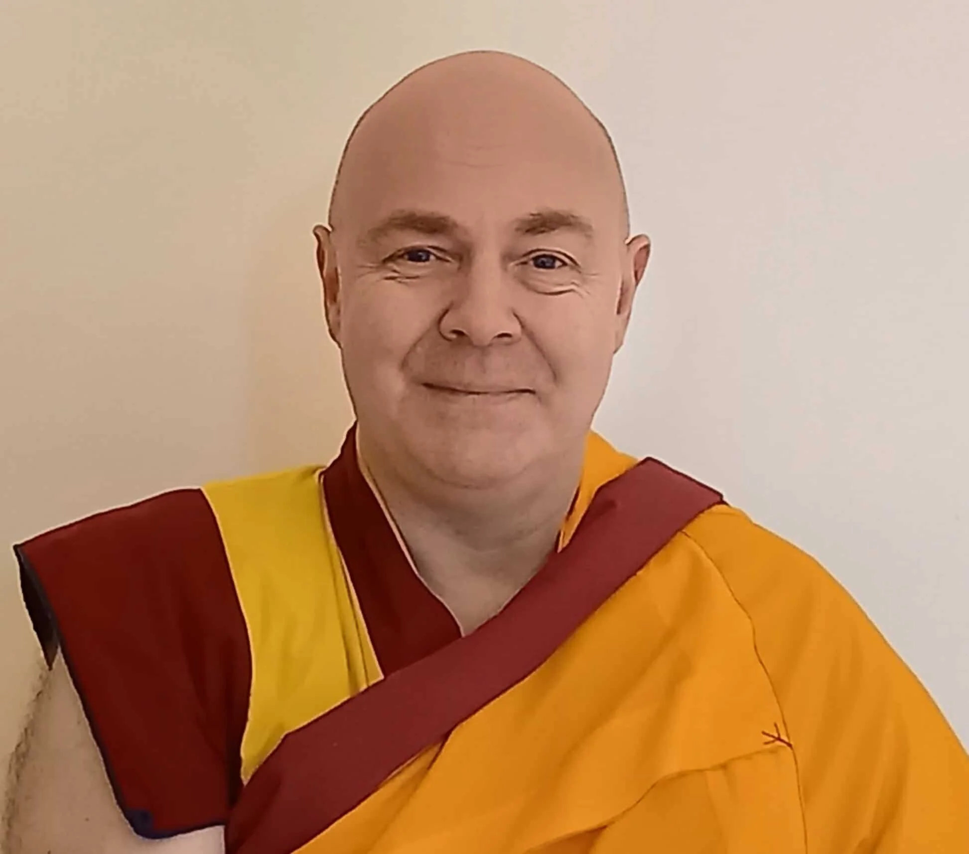 Resident Teacher, Gen Kelsang Jangchub, wearing maroon and yellow robes — a Buddhist monk, smiling against a plain light wall.