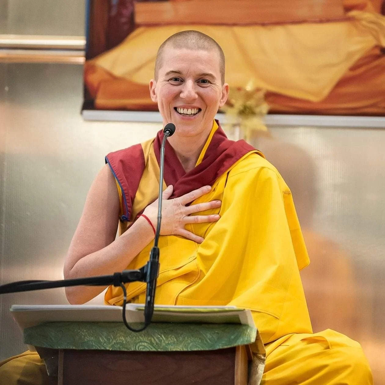 Buddhist nun Gen-la Kelsang sangden, Deputy General Spiritual Director of NKT-IKBU. She's smiling broadly, teaching with her hand on her heart, in front of a Kadampa Buddhist shrine with a photo of her teacher behind.