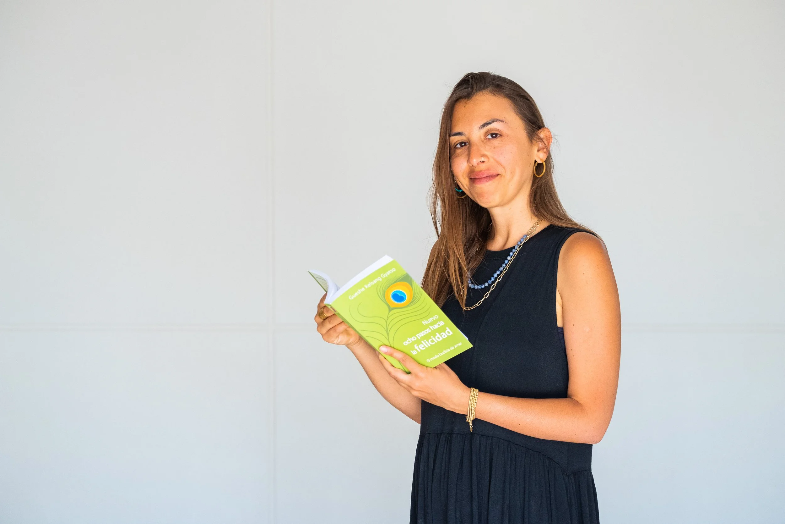 A young woman with long brown hair, wearing a black sleeveless dress is holding the Spanish language version of the book New eight Steps to Happiness with a peacock feather illustration on the cover.