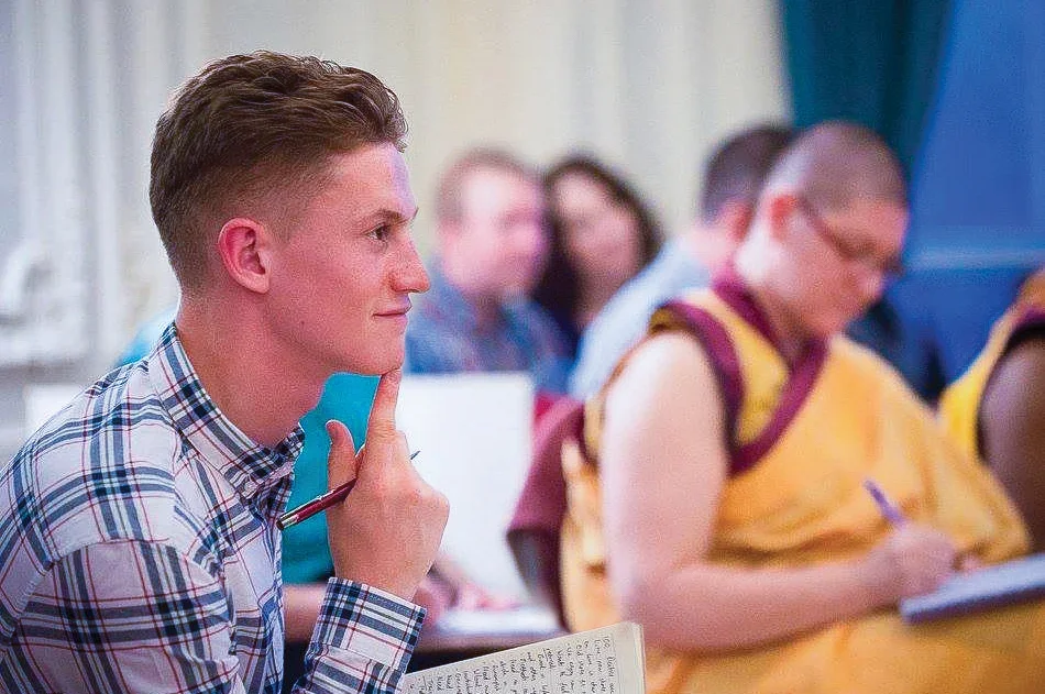 Young man with a checked shirt sitting in a classroom, with pen on his chin, listening attentively, with other students in the background.