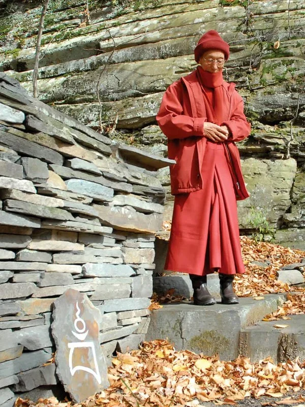 Venerable Geshe Kelsang Gyatso Rinpoche visiting the New York Kadampa World Peace Temple. He's dressed in a red coat and red hat walking down stone steps outdoors surrounded by fallen autumn leaves.