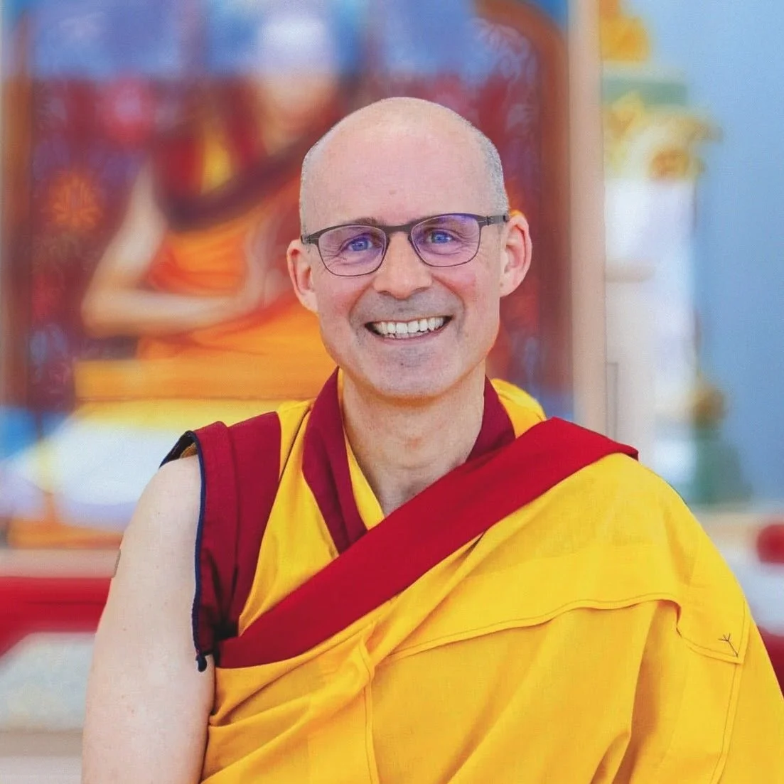 Buddhist monk Gen-la Kelsang Jampa, General Spiritual Director of NKT-IKBU. He's smiling, wearing glasses and teaching, in front of a Kadampa Buddhist shrine with a photo of his teacher behind.