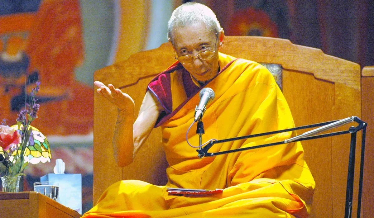 Venerable Geshe Kelsang Gyatso Rinpoche in US Kadampa temple teaching. He's wearing glasses, speaking into a microphone, with his hand raised in a gesture of offering the listener something.