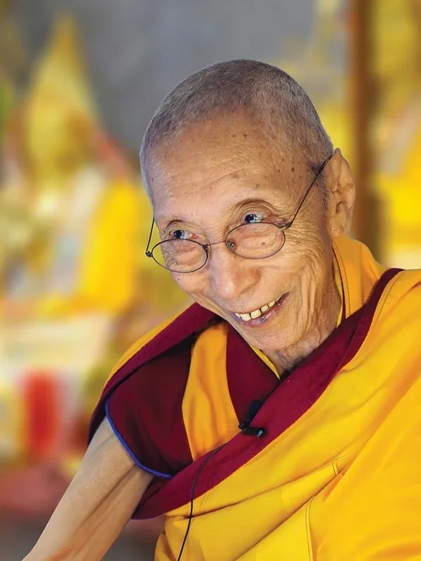 Venerable Geshe Kelsang Gyatso Rinpoche as an elderly Buddhist monk. He's teaching, leaning forwards, wearing glasses and smiling kindly.