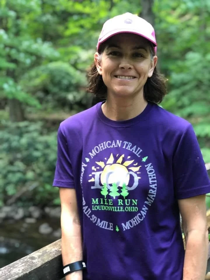 A woman smiling outdoors, wearing a purple MHCAN Trail T-shirt and a pink cap, standing near a wooded area with a stream.
