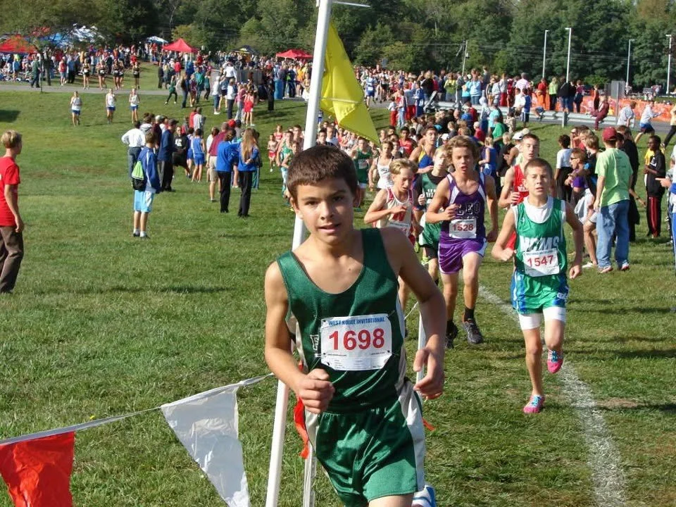 Young boys and girls competing in a cross-country race in a grassy park, surrounded by spectators and supporters.