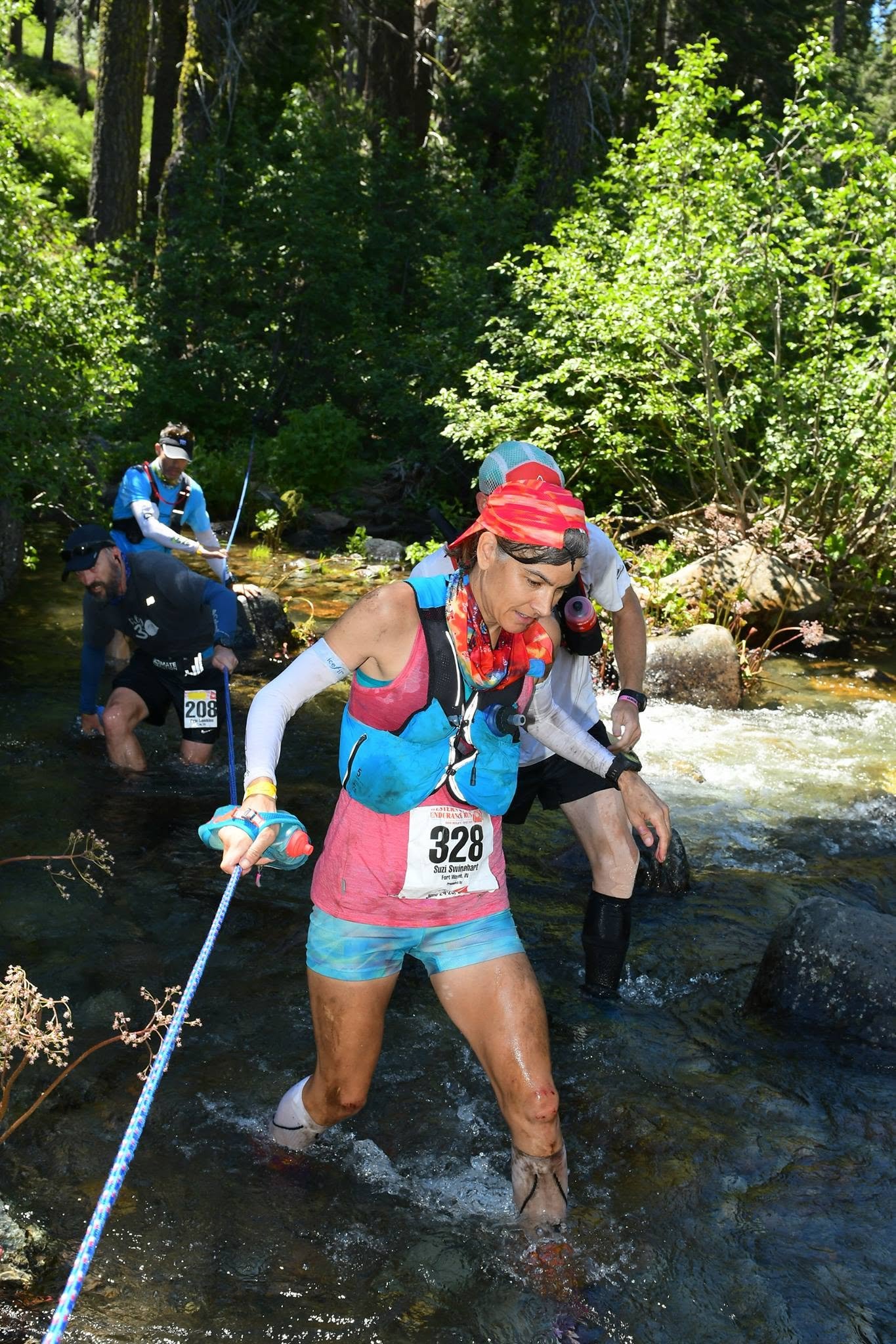 Group of runners crossing a shallow stream in a forest during a trail race.