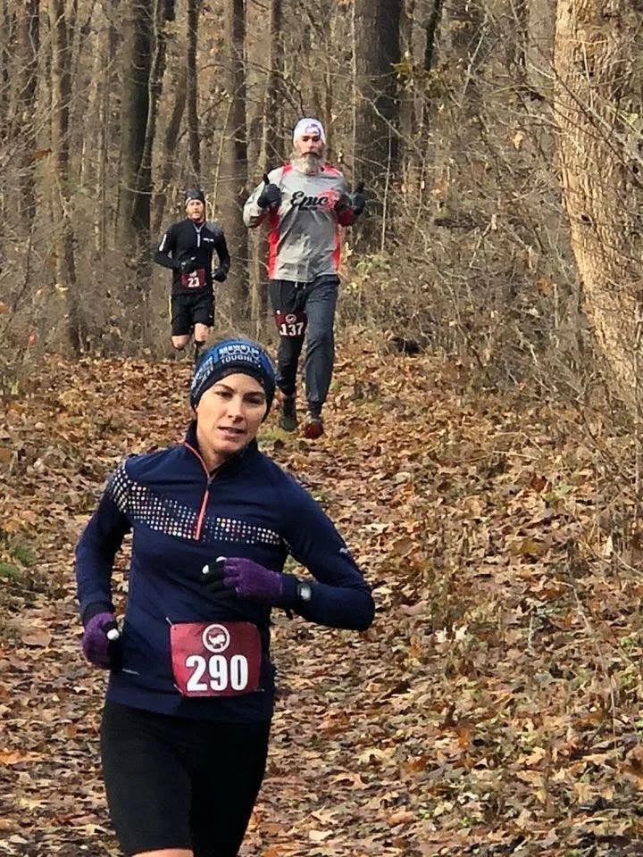 Three runners participating in a trail race through a wooded area with fallen autumn leaves.