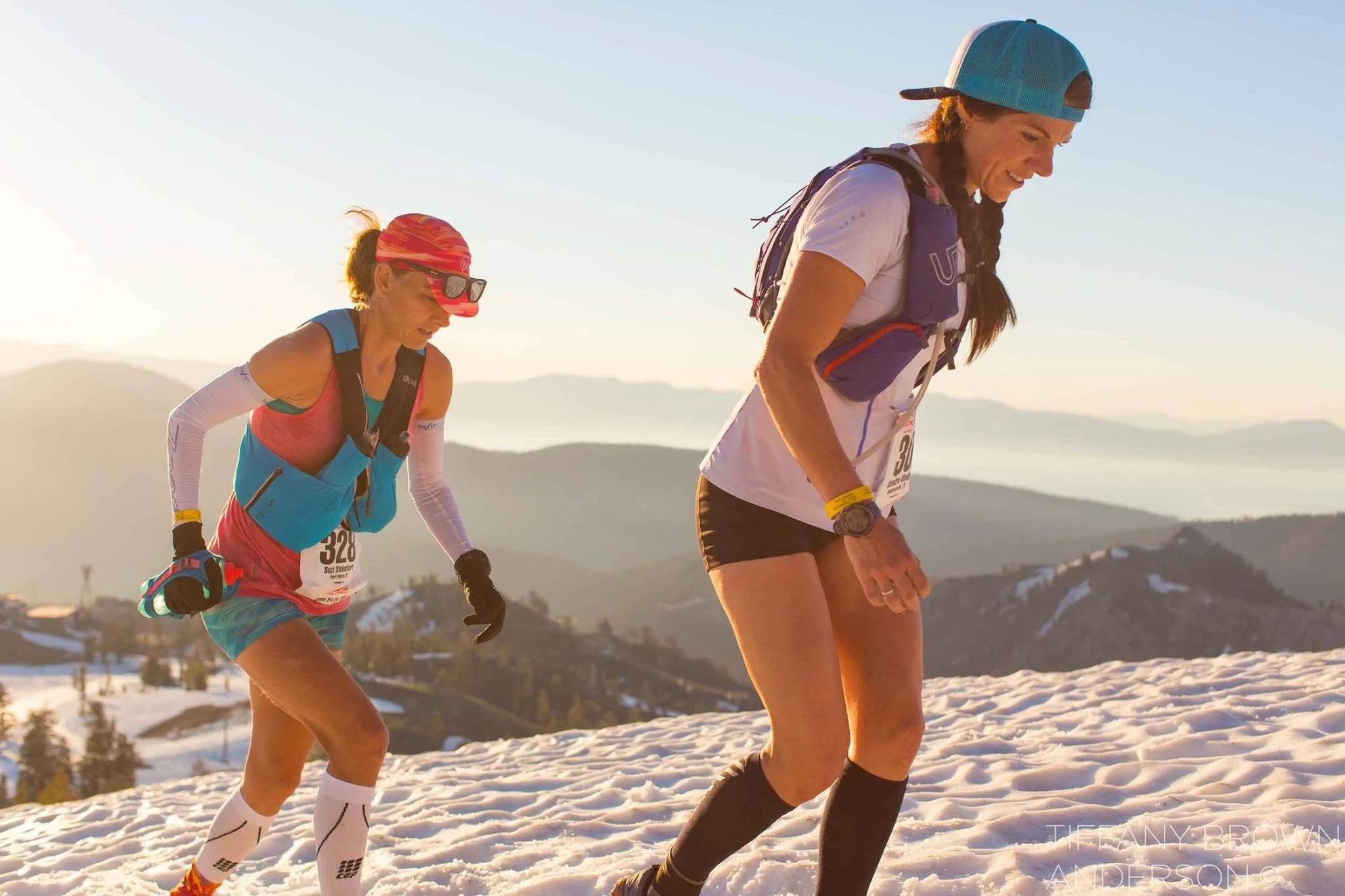Two women are running or hiking in a snowy landscape during sunset or sunrise, wearing athletic clothing, hats, and backpacks.