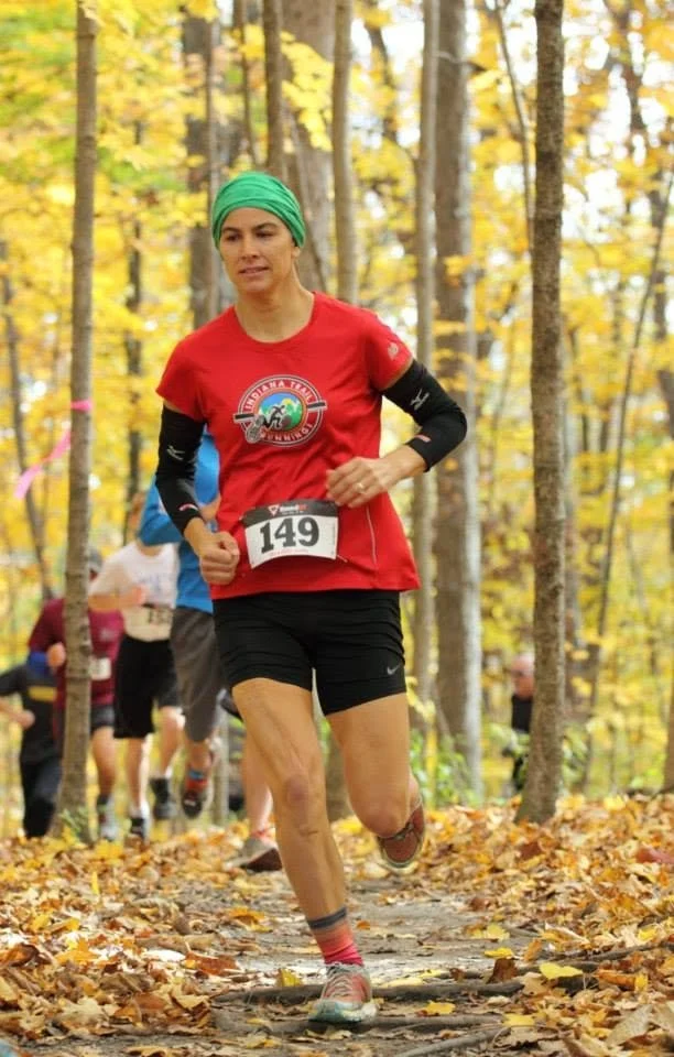 A woman running in a forest during autumn, wearing a red shirt, black shorts, a green headwrap, and a race bib number 149.