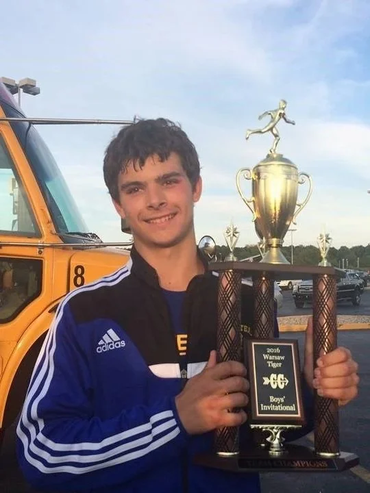 A young man in a blue and black Adidas jacket holding a trophy with a plaque that says "2016 Waraw Tiger Boys Invitional" and a picture of a tiger. Behind him is a yellow school bus and cars in a parking lot.