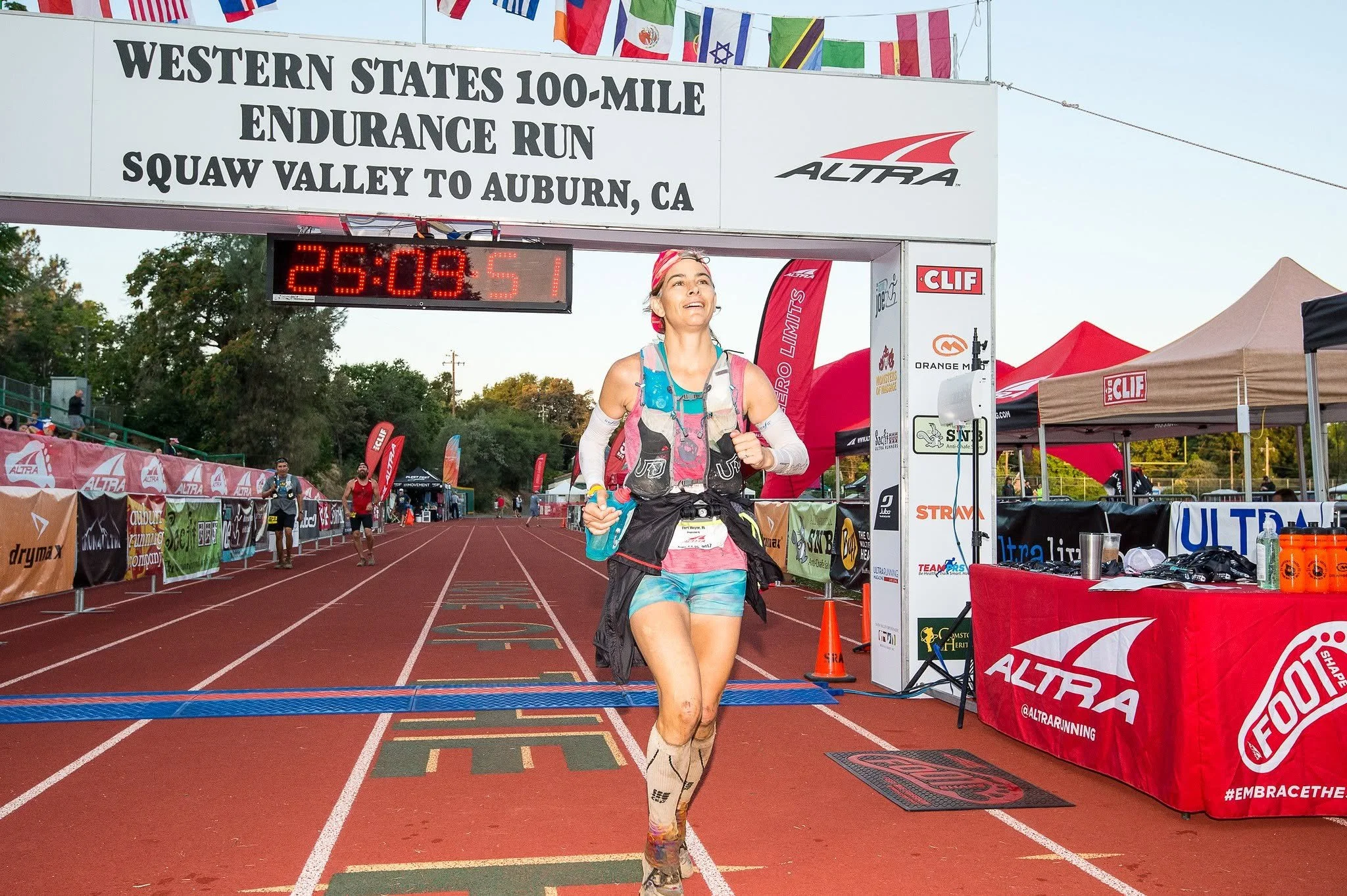 A woman running in a race at a stadium finish line, smiling as she crosses the finish line with a digital timer displaying 25:09:51, surrounded by banners, tents, and flags.