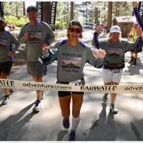 A group of people participating in a trail running event in a wooded area, with a woman crossing the finish line smiling and raising her arms in celebration.