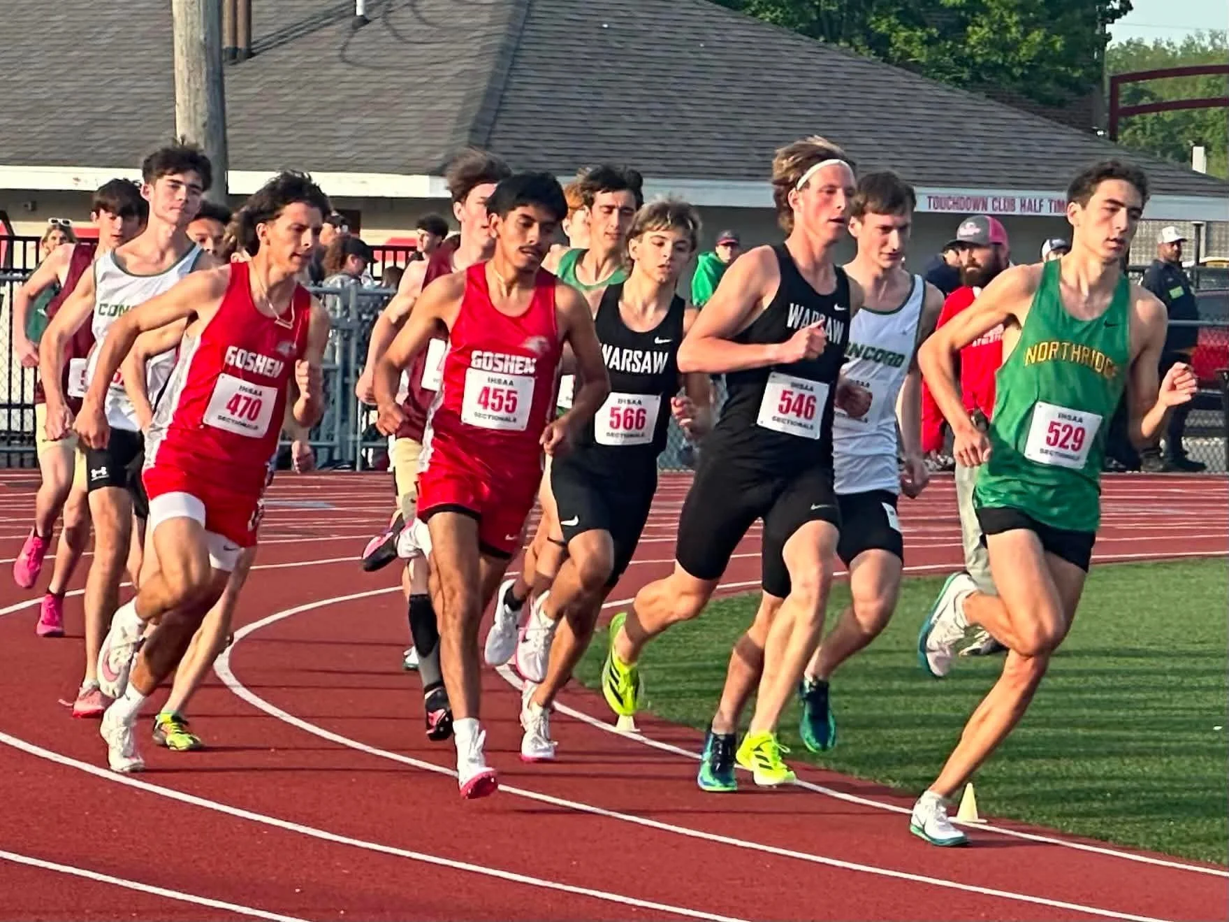 A group of young male athletes running on a red track during a race, wearing different athletic uniforms and race bibs.