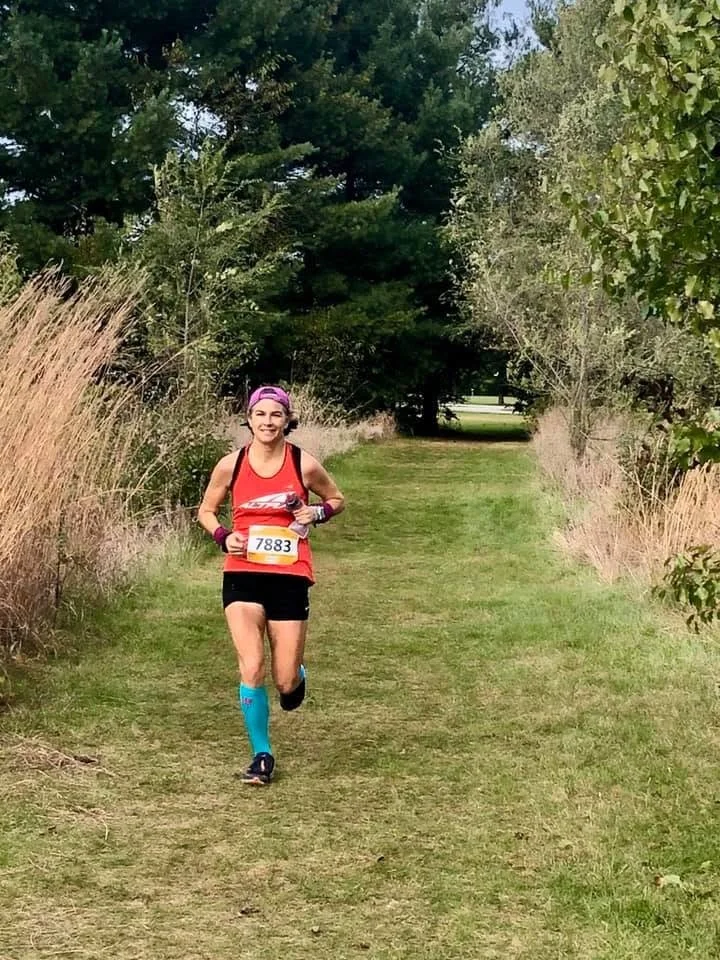 A woman running along a grassy trail surrounded by trees and tall grass, wearing a pink cap, red running top, black shorts, blue compression socks, and a race bib number 7883.