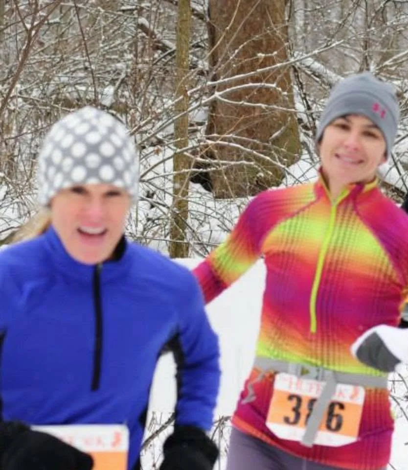 Two women in winter running attire smiling and jogging outdoors in a snowy forest.