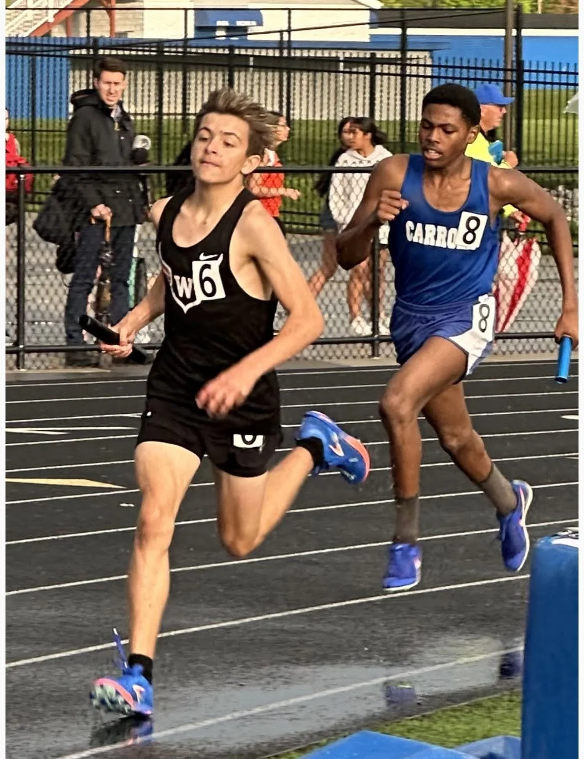 Two male runners competing in a track race, one in a black uniform with the number 6 and the other in a blue uniform with the number 8, with a fence and spectators in the background.