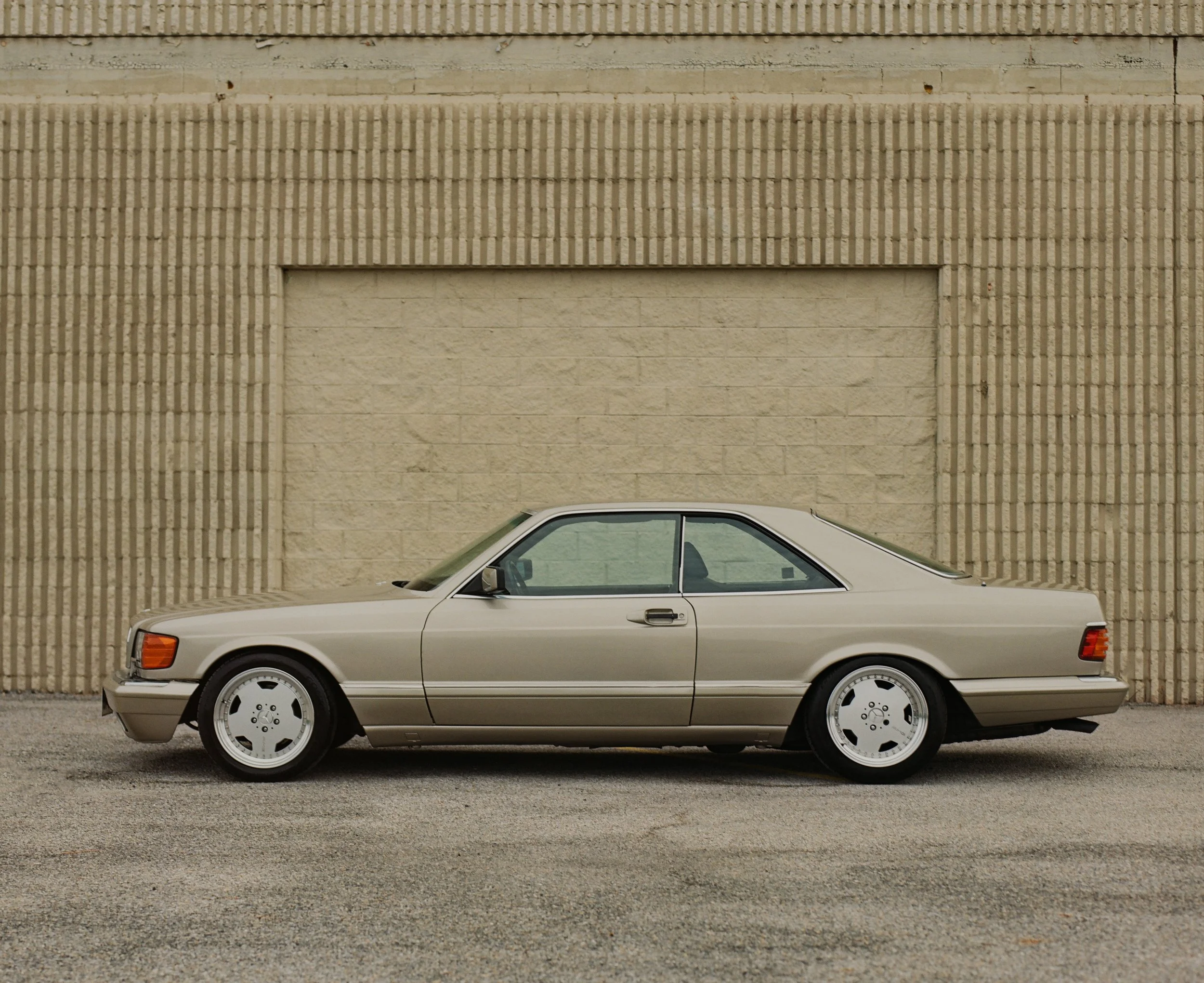A vintage beige car parked in front of a beige brick wall with a small window near the center. The car is positioned sideways, showing its side profile.