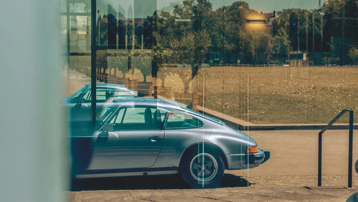 A vintage silver sports car parked outside a glass building on a sunny day, with trees and a grassy area in the background.
