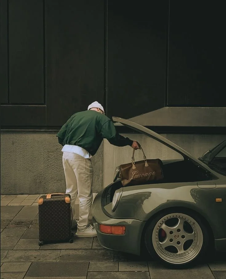 A man with a suitcase and a brown bag opening the hood of a dark gray vintage Porsche 911 parked on the street.