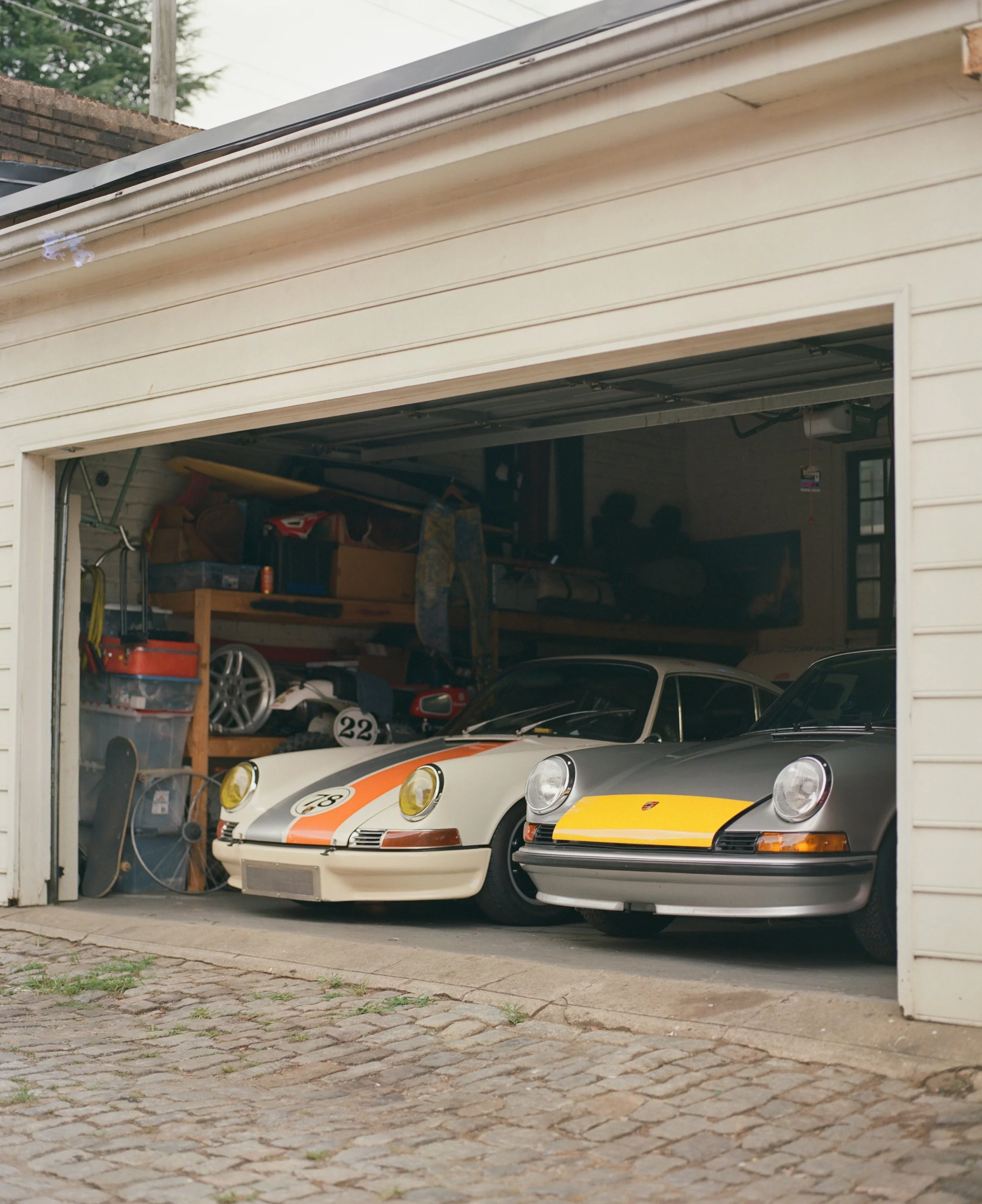 Garage with two classic Porsche cars parked inside, one white with orange racing stripes and the other gray with yellow accents. The garage is cluttered with various items and tools, and the driveway is made of cobblestone.