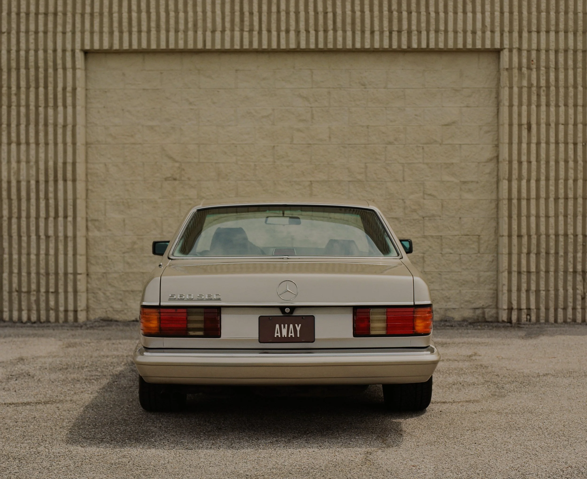 A vintage white Mercedes-Benz 300SEC car parked in front of a beige brick wall with vertical grooves. The car's rear is visible, with a black license plate that reads "AWAY." The car features classic rectangular tail lights with amber, red, and white sections.