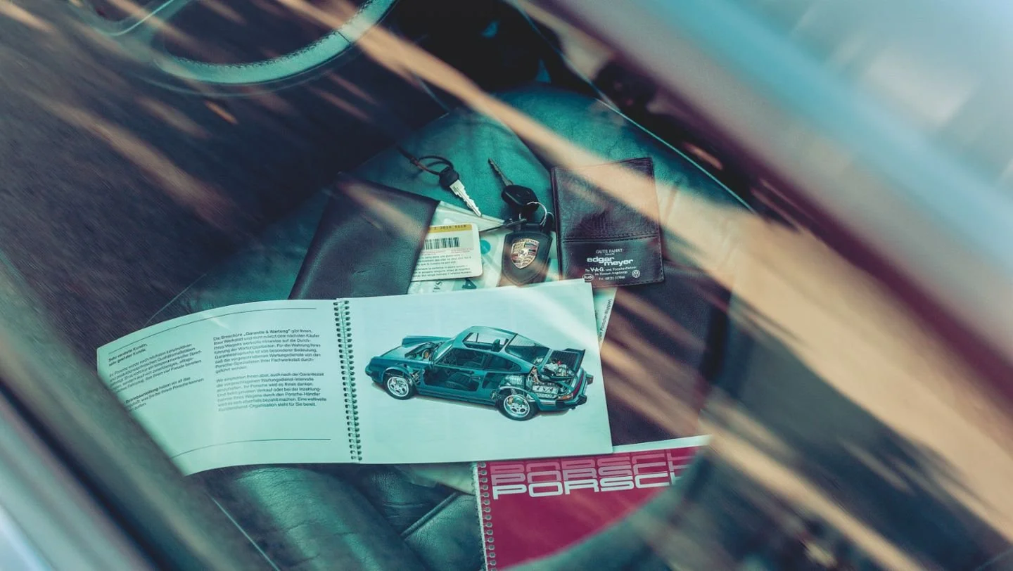 Open car manual showing a diagram of a Porsche car's interior and mechanical parts, placed on the dashboard with a Porsche brochure underneath, alongside car keys and identification cards on a black leather surface.