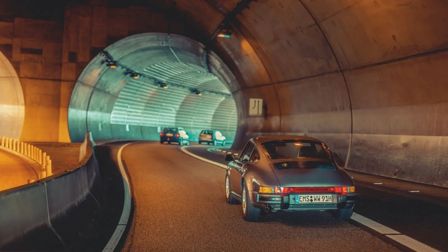 A vintage sports car driving through a tunnel with other cars ahead, illuminated by tunnel lights.