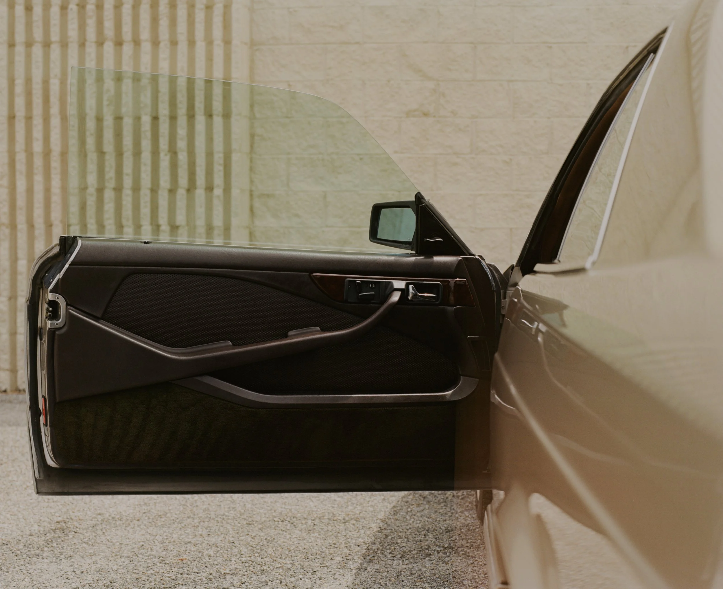 A close-up of a car door with the side mirror visible, and a clear windshield with a partial view of the interior, set against a yellow brick wall and a beige wall.