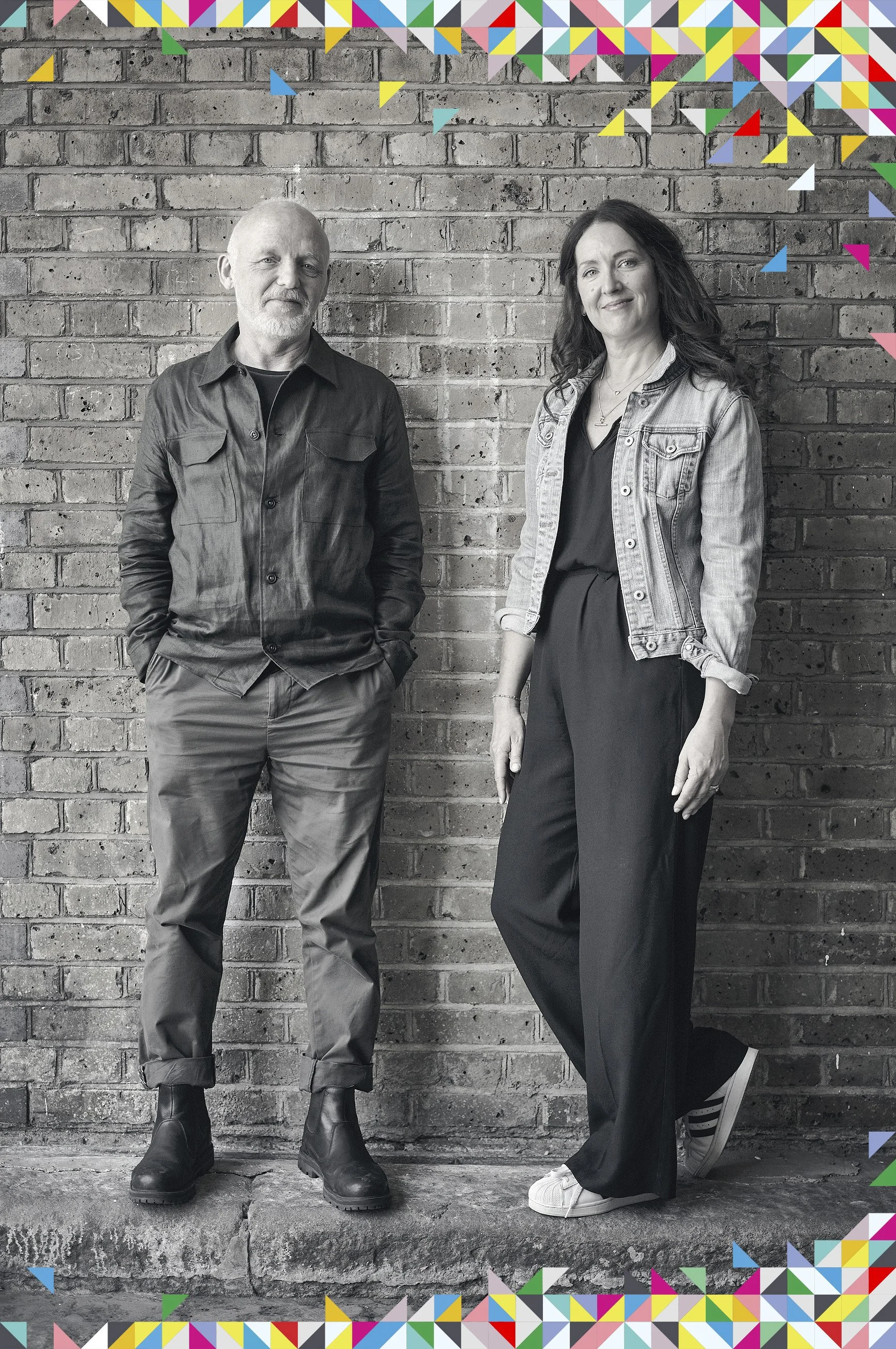 Black and white photo of a man and woman standing on a concrete step in front of a brick wall. The man wearing a long-sleeved button-up shirt and pants. The woman has long dark hair, wearing a denim jacket over a dark outfit and white sneakers. Colorful triangular border pattern surrounds the photo.