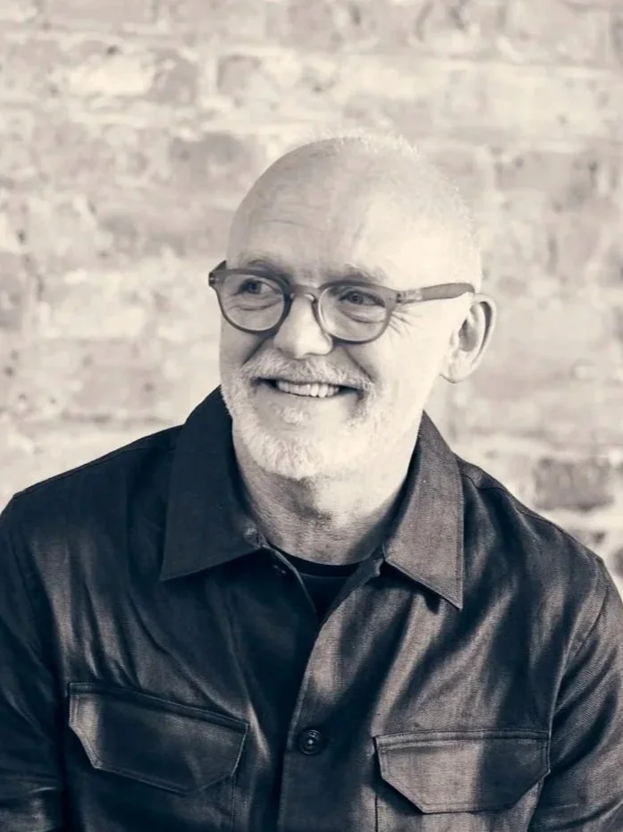 Black and white photo of an older man with glasses, a beard, and a mustache, smiling, wearing a leather jacket.