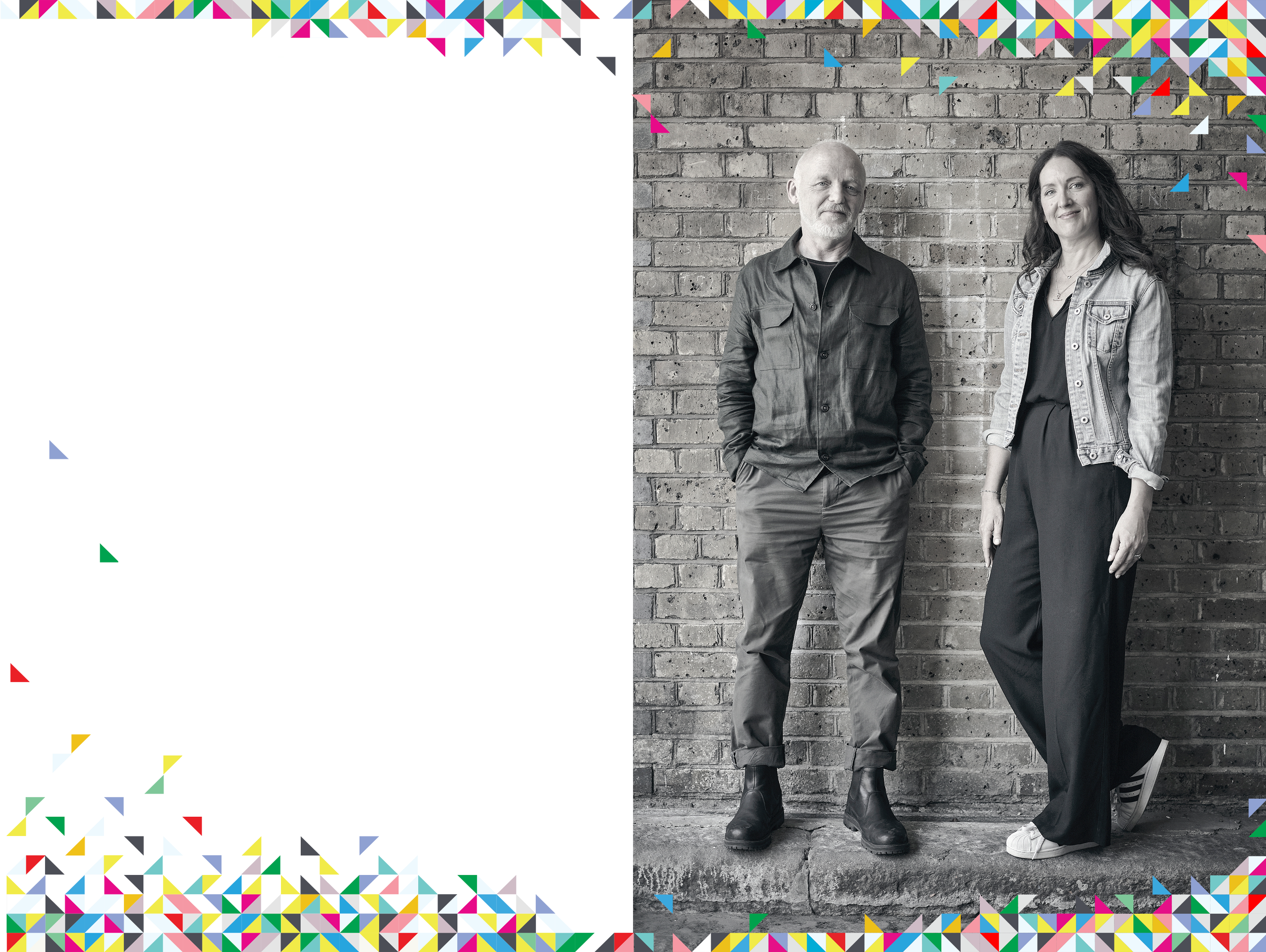 Black and white photo of a man and woman standing against a brick wall. The man has a beard, and is wearing a button-up shirt and pants. The woman has long, wavy hair, and is dressed in a denim jacket over a black top and pants. They are smiling.