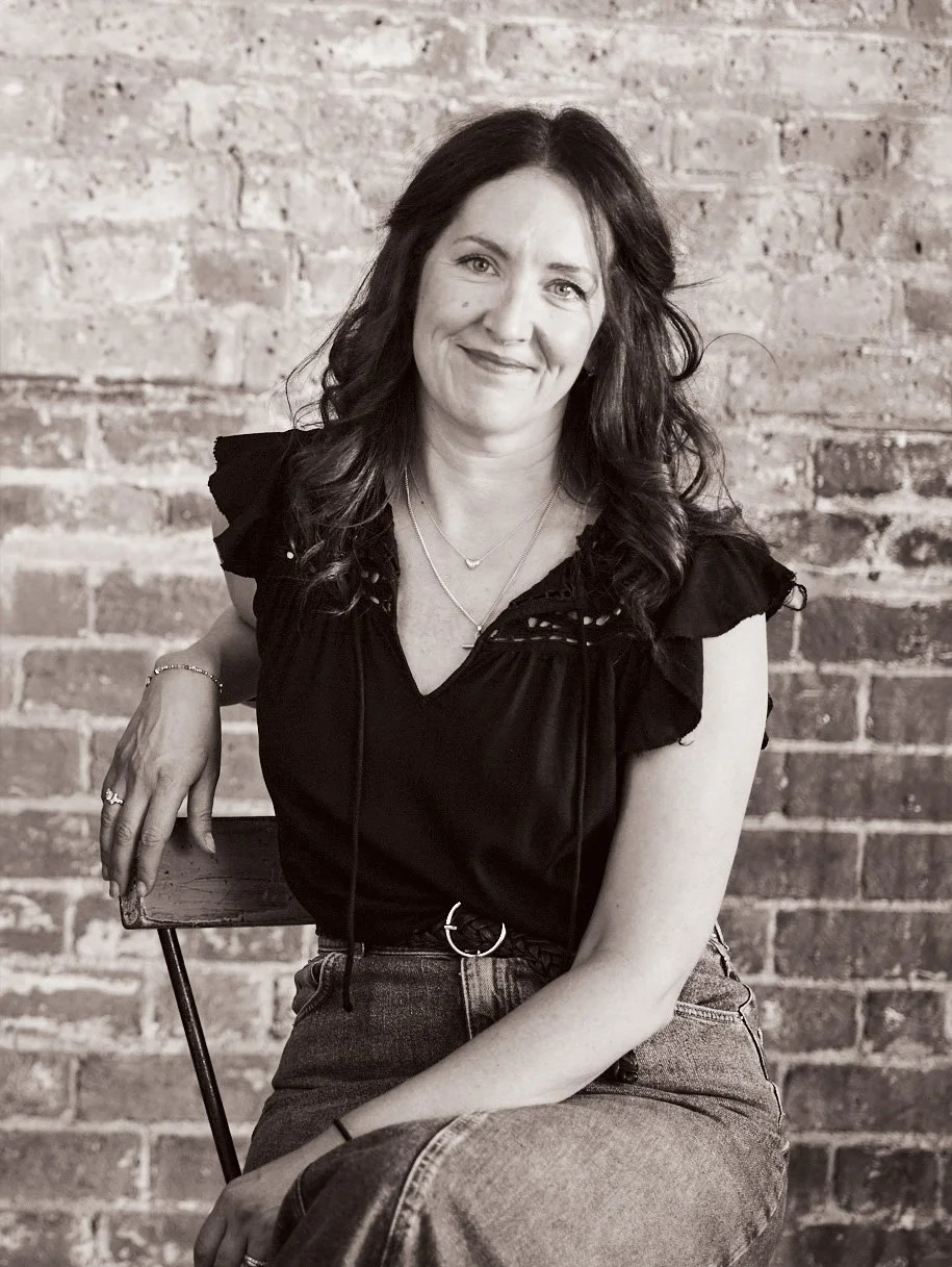A woman with dark, wavy hair and light skin sitting on a wooden chair, smiling at the camera, in front of a brick wall background.