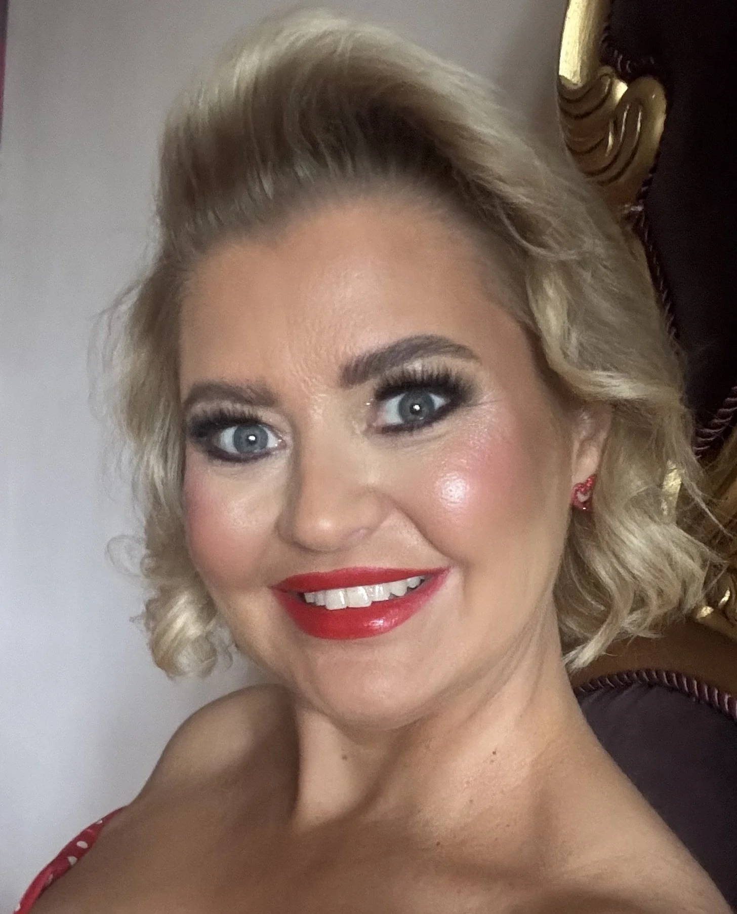 A smiling woman with blonde curly hair and blue eyes, wearing bright red lipstick and a red polka-dot dress, sitting on a dark-colored chair with gold accents.