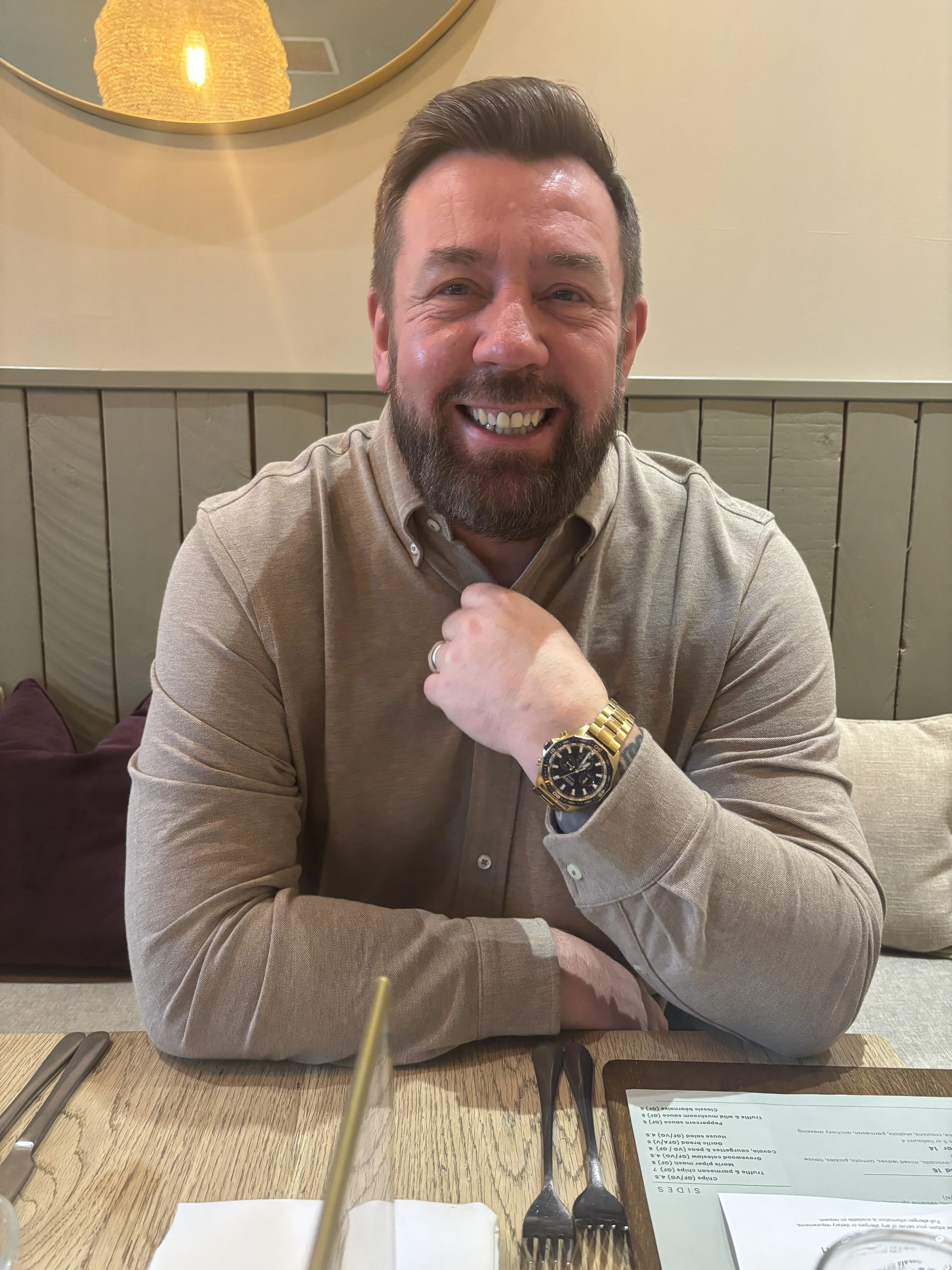 A man with dark hair and a beard, smiling and seated at a restaurant table. He's wearing a beige button-up shirt and a gold wristwatch. There is a mirror on the wall behind him with a warm light reflection.