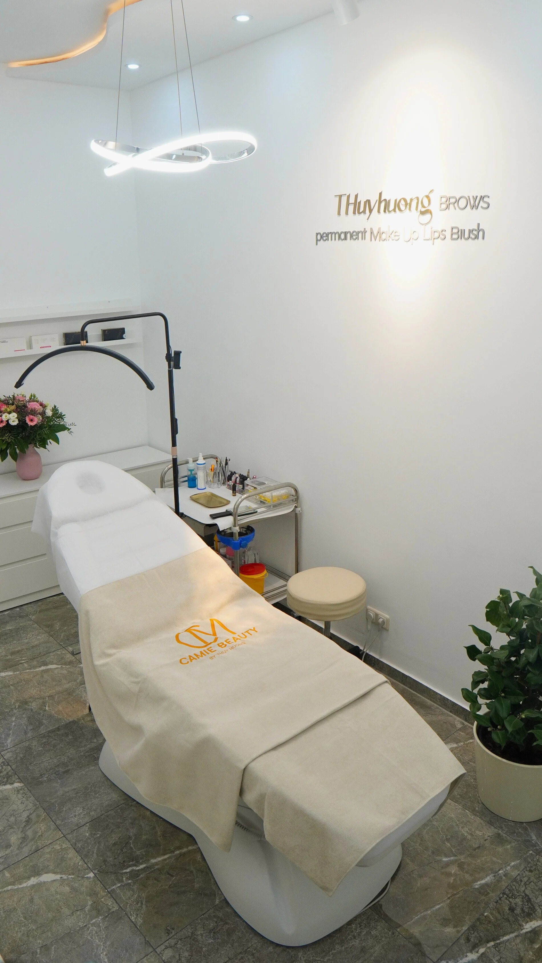 Ein Behandlungsstuhl in einem Kosmetikstudio mit einem Tuch darauf, daneben Kosmetikprodukte und Zubehör auf einem kleinen Tisch, im Hintergrund ein Blumenstrauß und Beratungssessel, an der Wand steht ein digitaler Beleuchtungskreis. Es ist ein moderner Raum für Schönheitsbehandlungen.