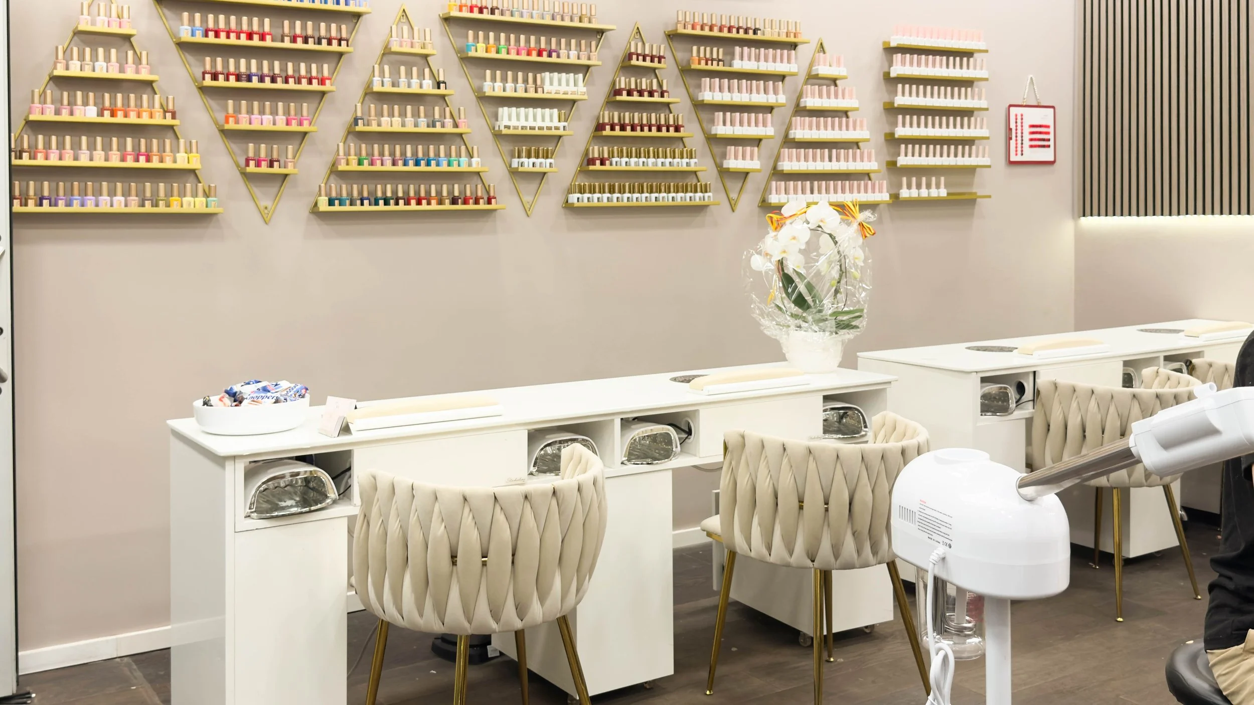 Beauty-Salon mit Gold im Hintergrund an den Wänden, weiße Tische mit Näh- oder Nagelzubehör, beige gepolsterte Stühle, eine Vase mit weißen Blumen auf einem Tisch, Geräte für Maniküre oder Pediküre.