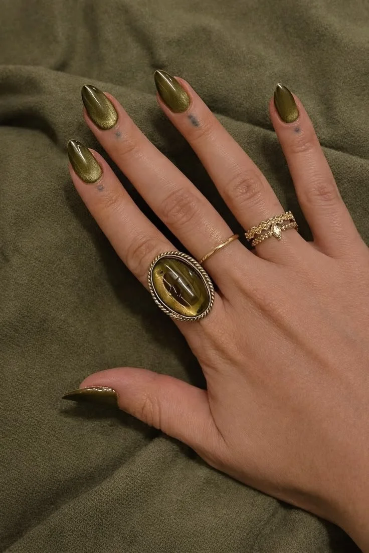 Nahaufnahme einer Hand mit kunstvollen, goldgrünen Nägeln, mehreren Ringen, darunter ein großer, ovaler Ring mit grünem Stein.