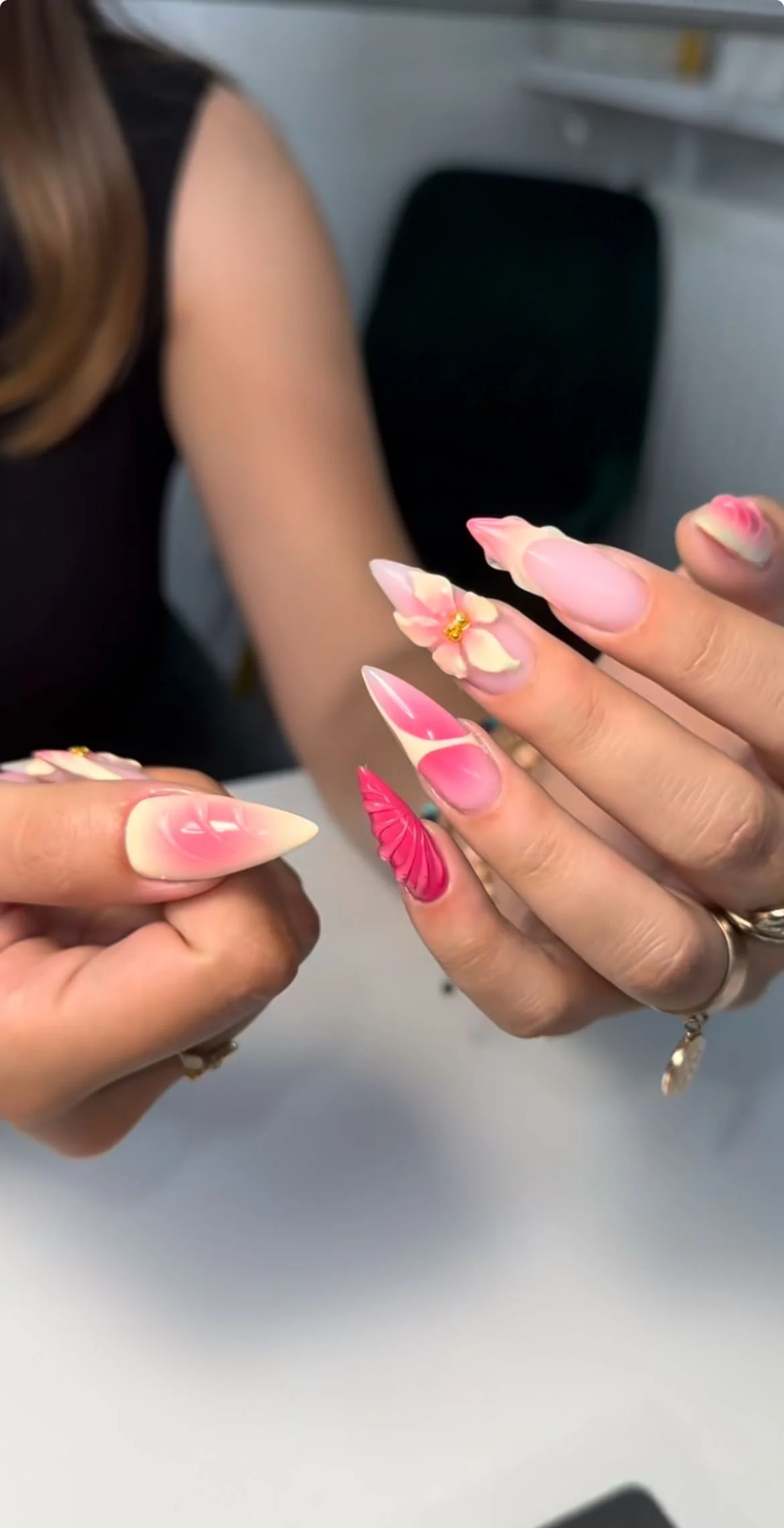 Nahaufnahme von Fingern mit kunstvollen, pink-weißen Nageldesigns, einem kleinen goldenen Dekor auf einem Nagel, und einem pinkfarbenen Nagel mit Muster. Eine Person zeigt ihre Hände, im Hintergrund ein schwarzer Stuhl.