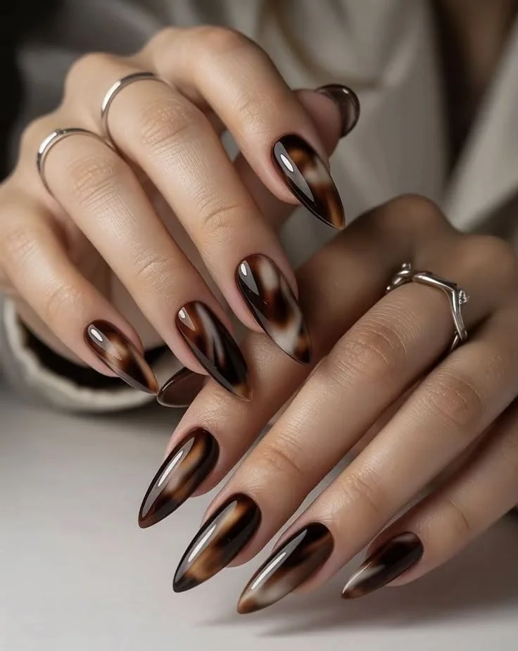 Nahaufnahme von manicürten Fingernägeln mit braun-schwarz-weißer Tiertarnung am Nagelrand, getragen auf einer Hand mit Ringen.