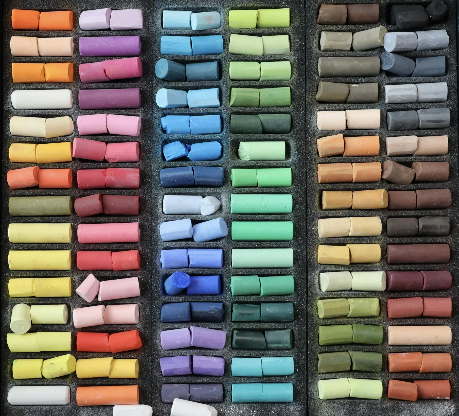A collection of colorful pastel sticks arranged in rows and organized by color, including shades of orange, purple, blue, green, beige, white, yellow, and gray, with some chalks showing signs of use.