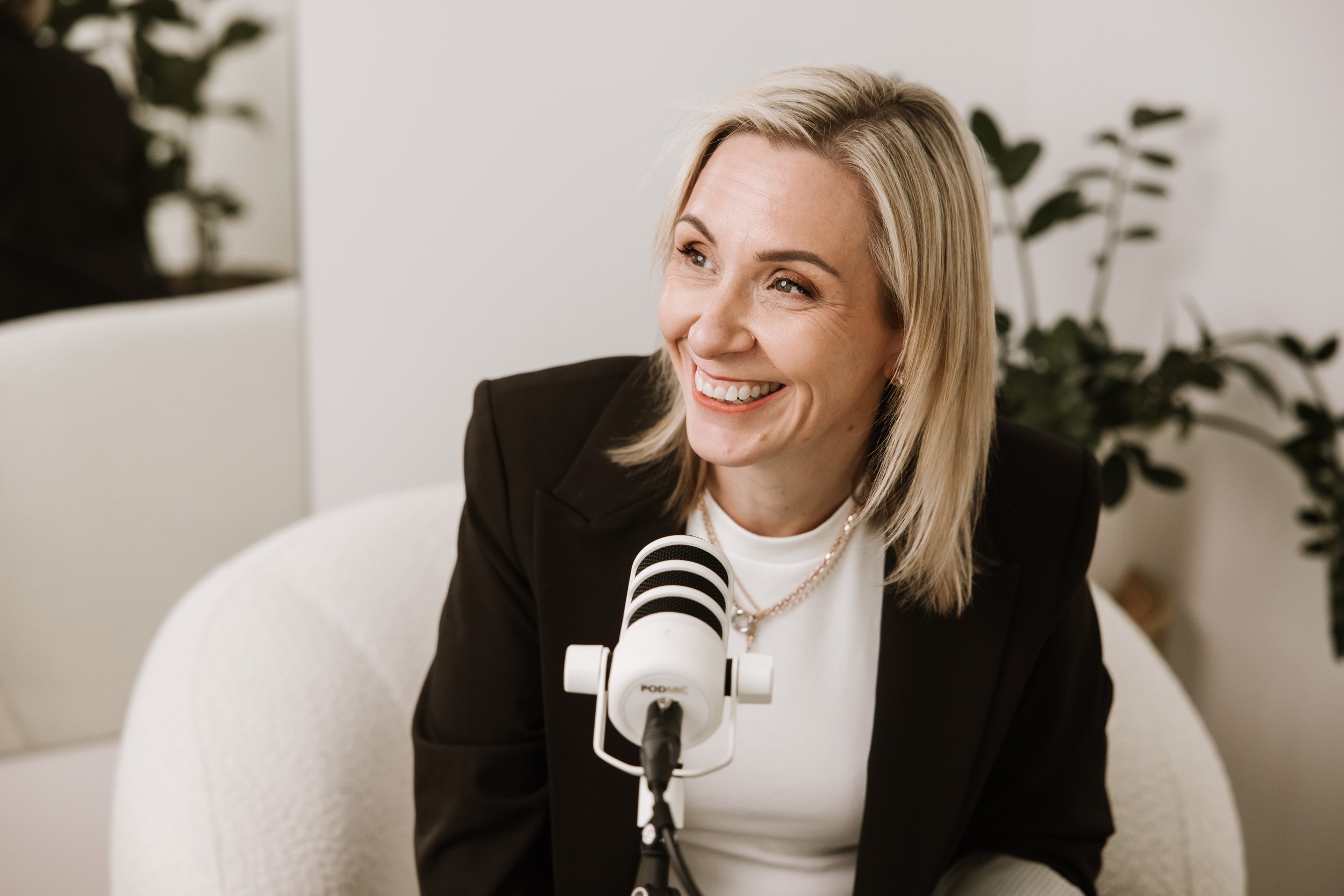 A smiling woman with blonde hair, wearing a black blazer and white shirt, speaking into a microphone during a podcast or interview, with a blurred plant in the background.