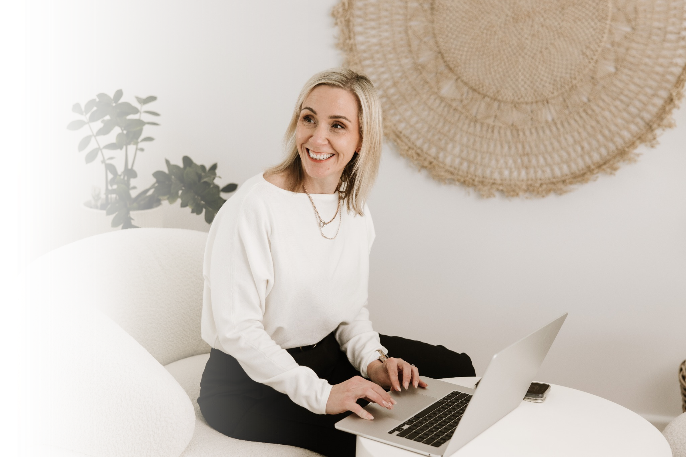 A woman with blonde hair, wearing a white shirt, sitting on a light-colored sofa with a laptop on her lap, smiling and typing. There is a potted plant and a large woven wall hanging in the background.
