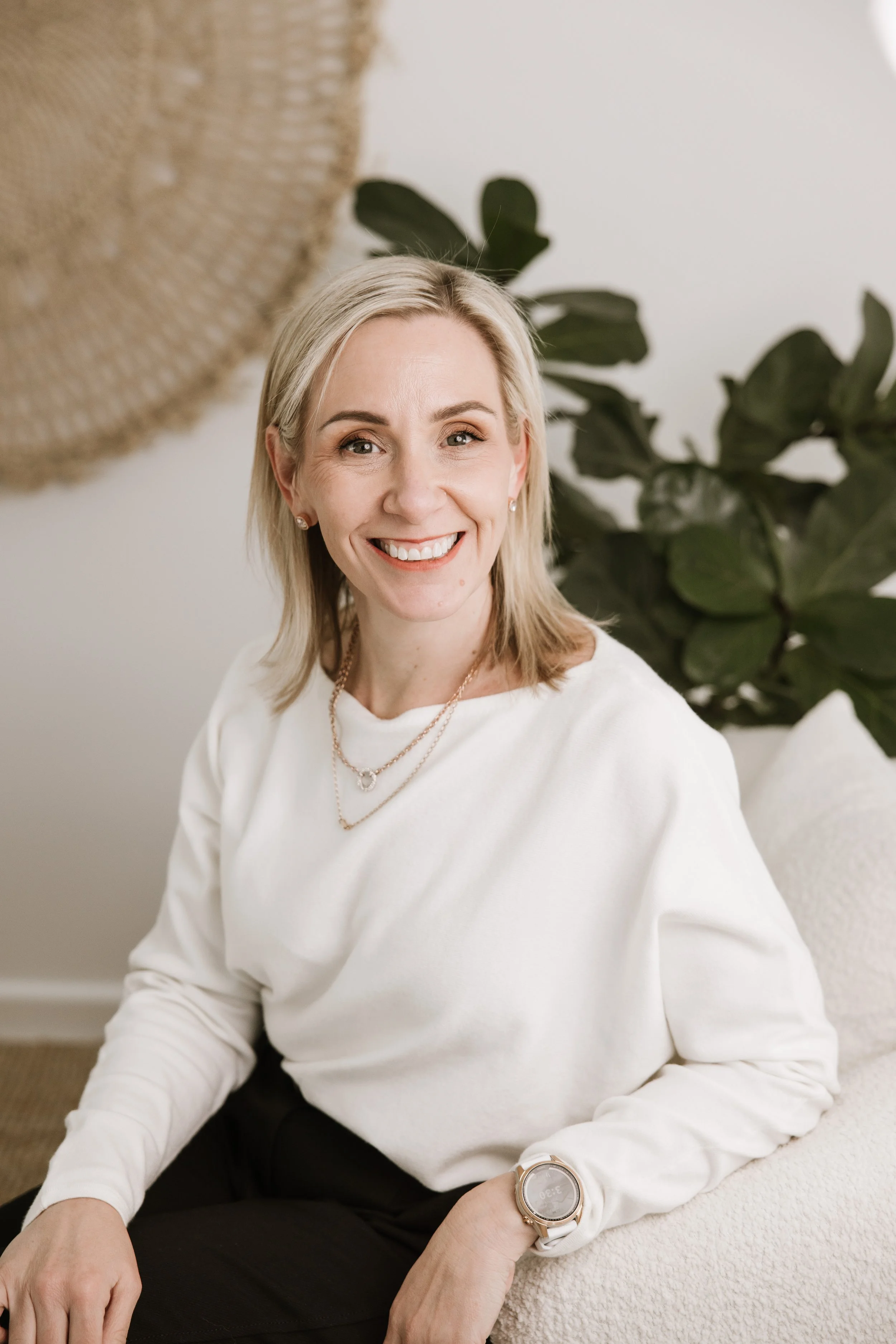A woman with blonde hair smiling while sitting on a white couch, wearing a white sweater, black pants, a silver watch, and jewelry, with a large green plant behind her.