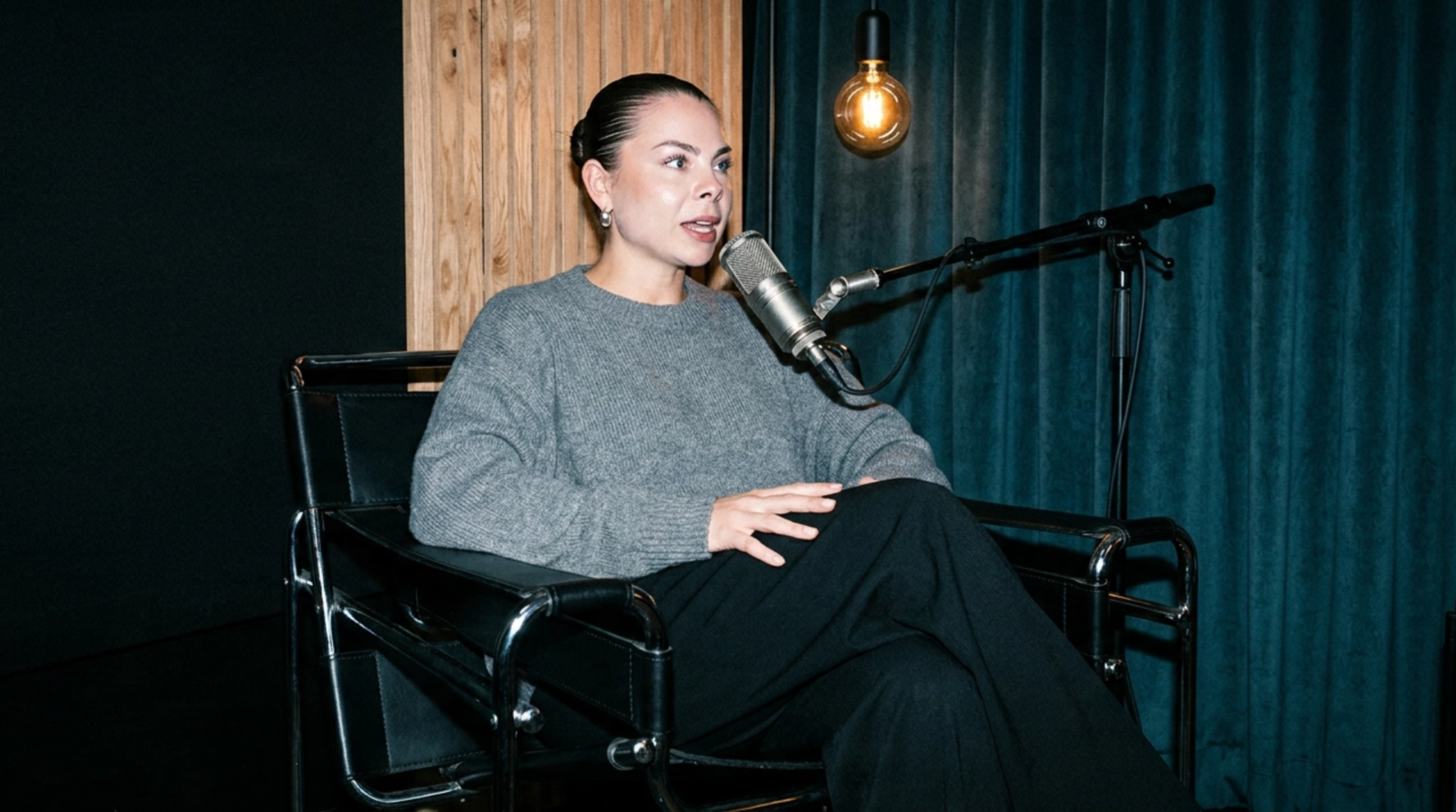 A woman with dark hair pulled back, wearing a gray sweater, sitting in a black chair with her legs crossed, speaking into a microphone, in a room with wooden and blue curtains.