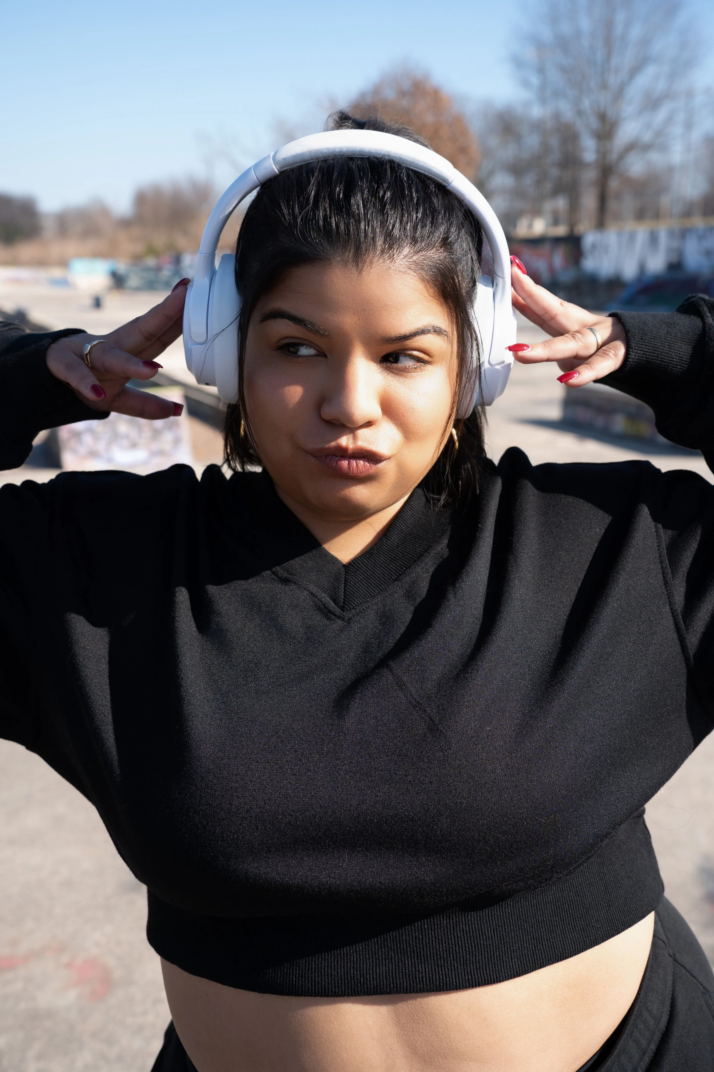A woman wearing large white headphones poses outdoors on a sunny day. She has dark hair pulled back, and is wearing a black hoodie. Her hand is raised to the headphones, and she makes a playful expression with puckered lips and slightly rolled eyes. The background shows bare trees and a graffiti-covered wall.